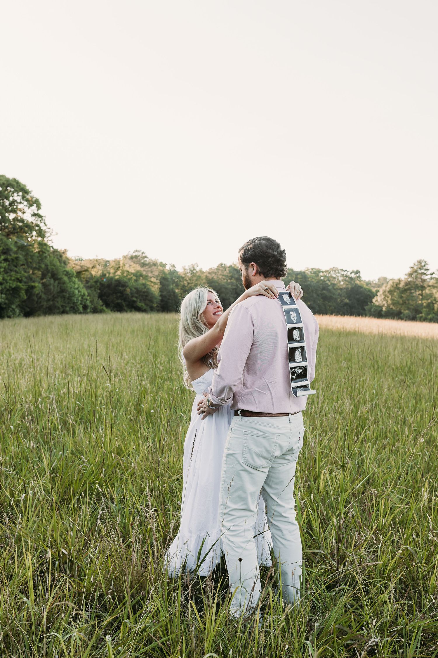 Couple embracing in a green field, man holding ultrasound photos behind his back during a maternity announcement shoot.
