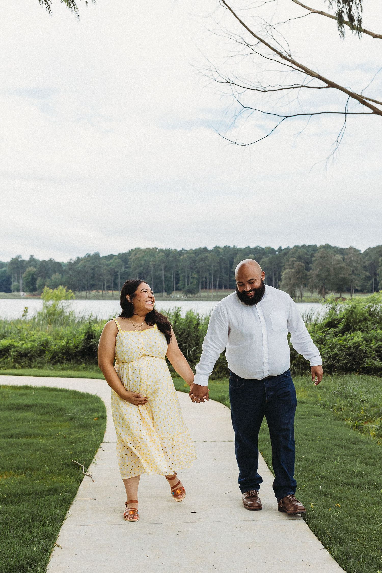 Pregnant woman in yellow dress holds hands with bearded man in white shirt walking on park path by lake.