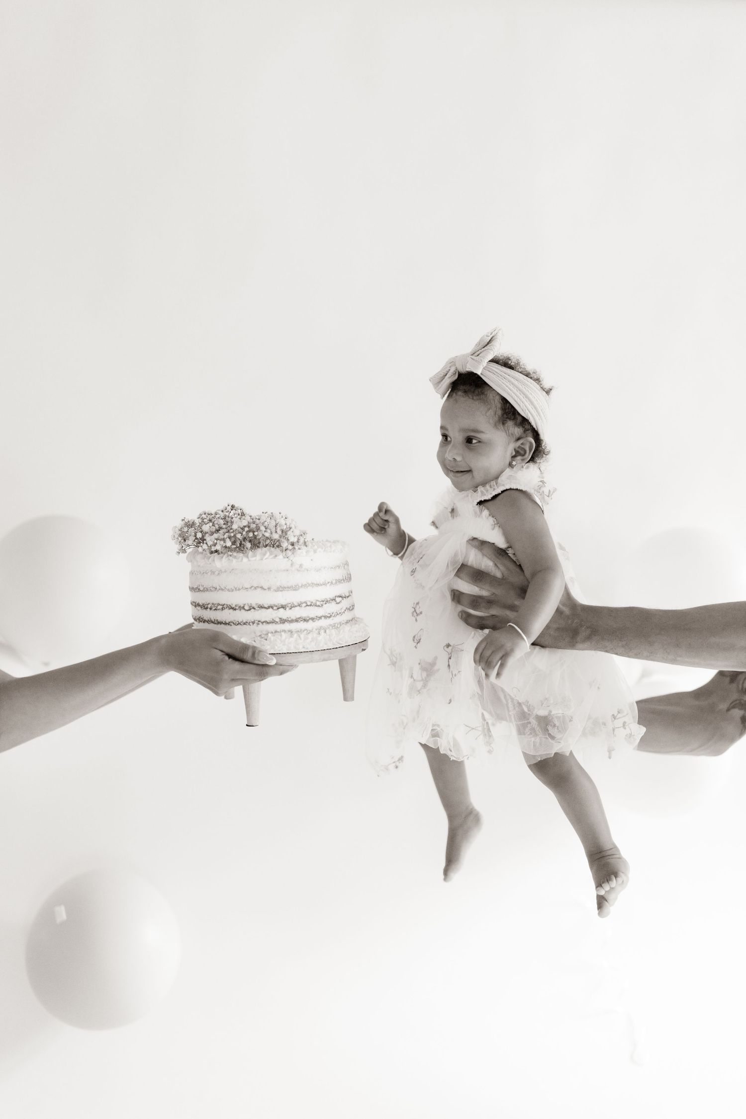 Black and white photo of a baby girl in white dress held by adult hands near a decorated birthday cake with balloons.