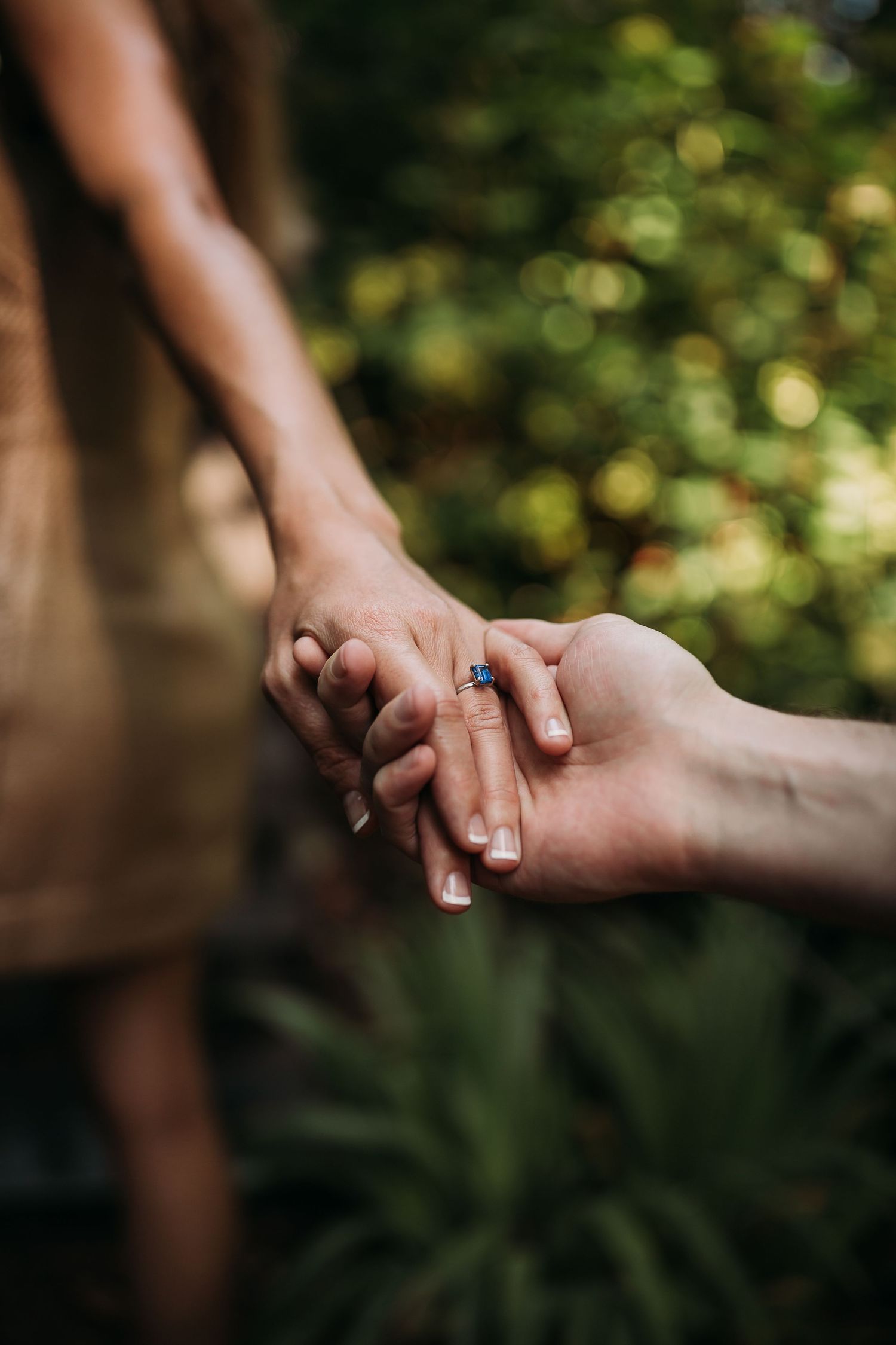 Close-up of couple holding hands outdoors, bride wearing a blue sapphire engagement ring.