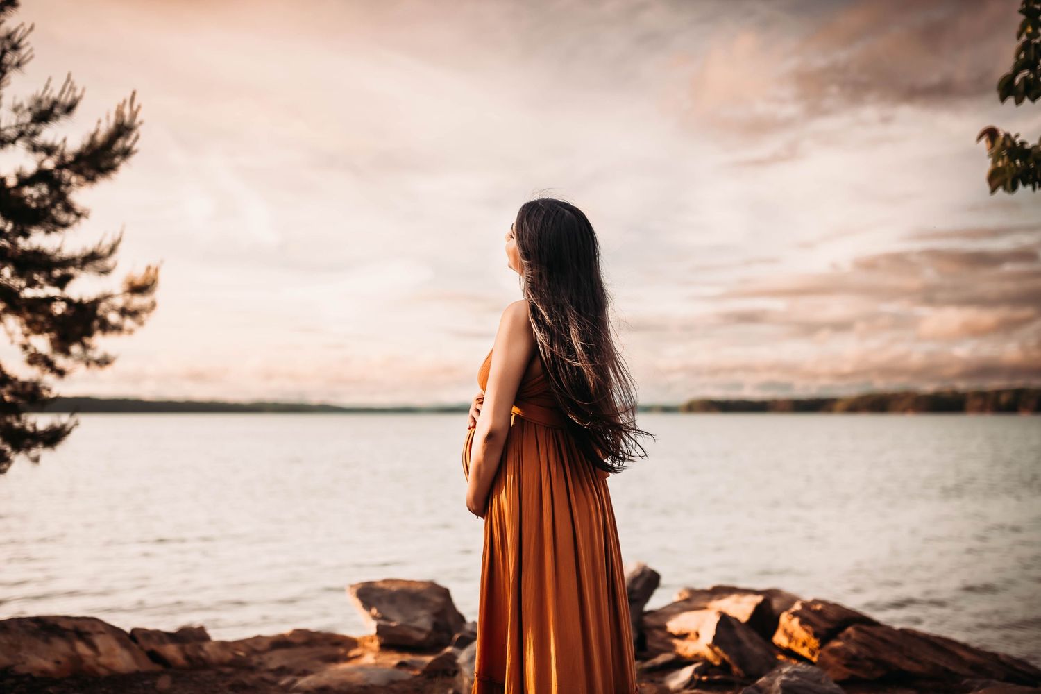 Woman in orange dress standing on rocky shore gazing at a serene lake during a golden sunset.