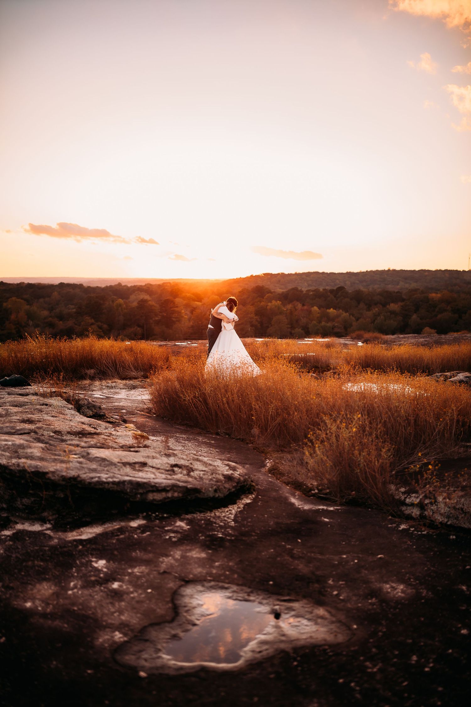 A bride in a white gown stands alone on rocky terrain at sunset with a glowing orange sky backdrop.