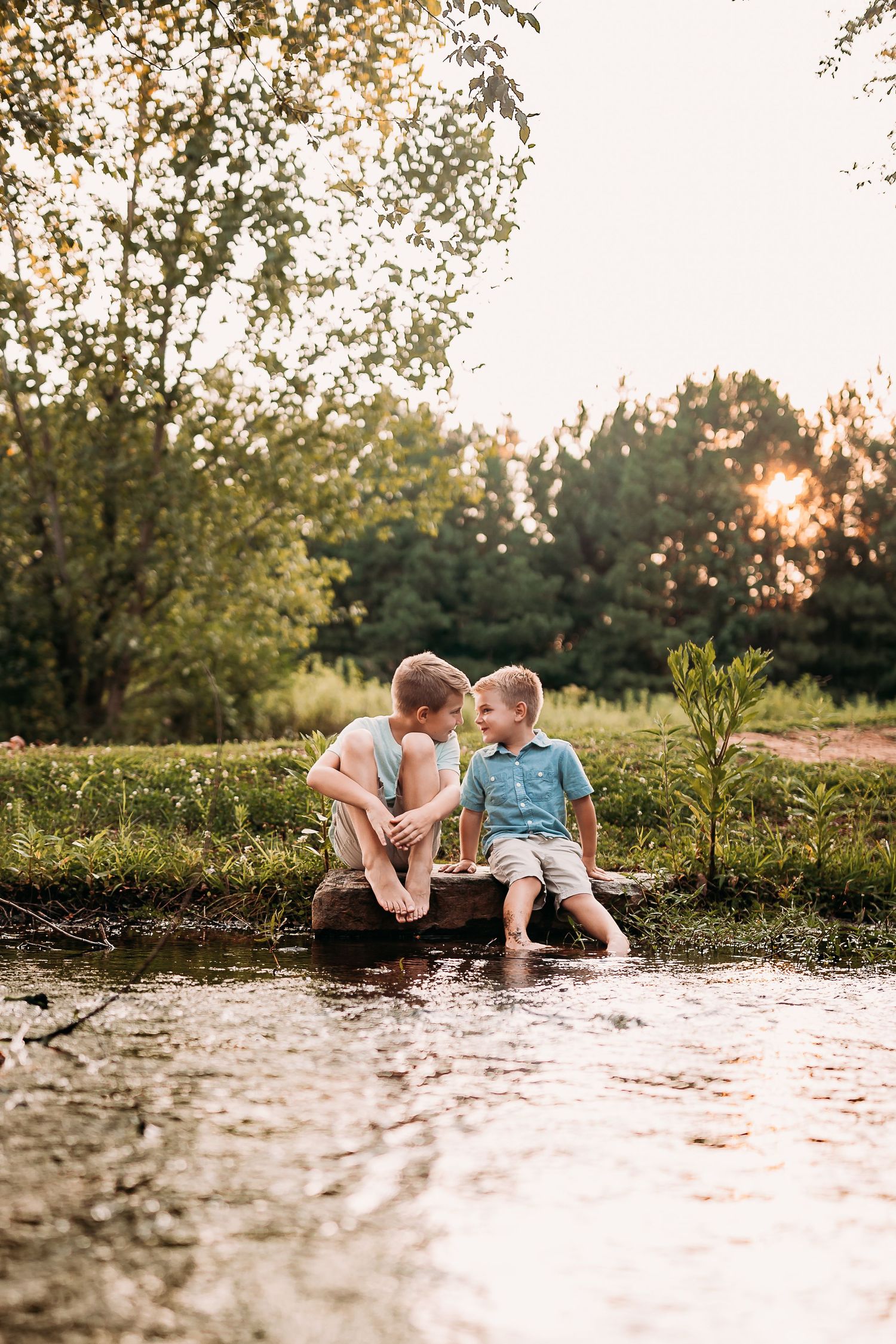 Two children sitting on a log by a peaceful pond, enjoying a sunset conversation surrounded by lush green trees.