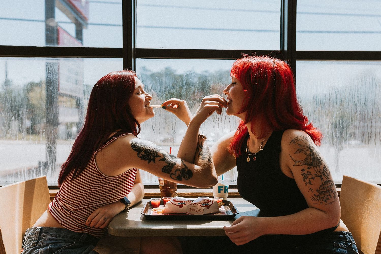 Two tattooed women with red hair sharing food and laughing together at a restaurant table.