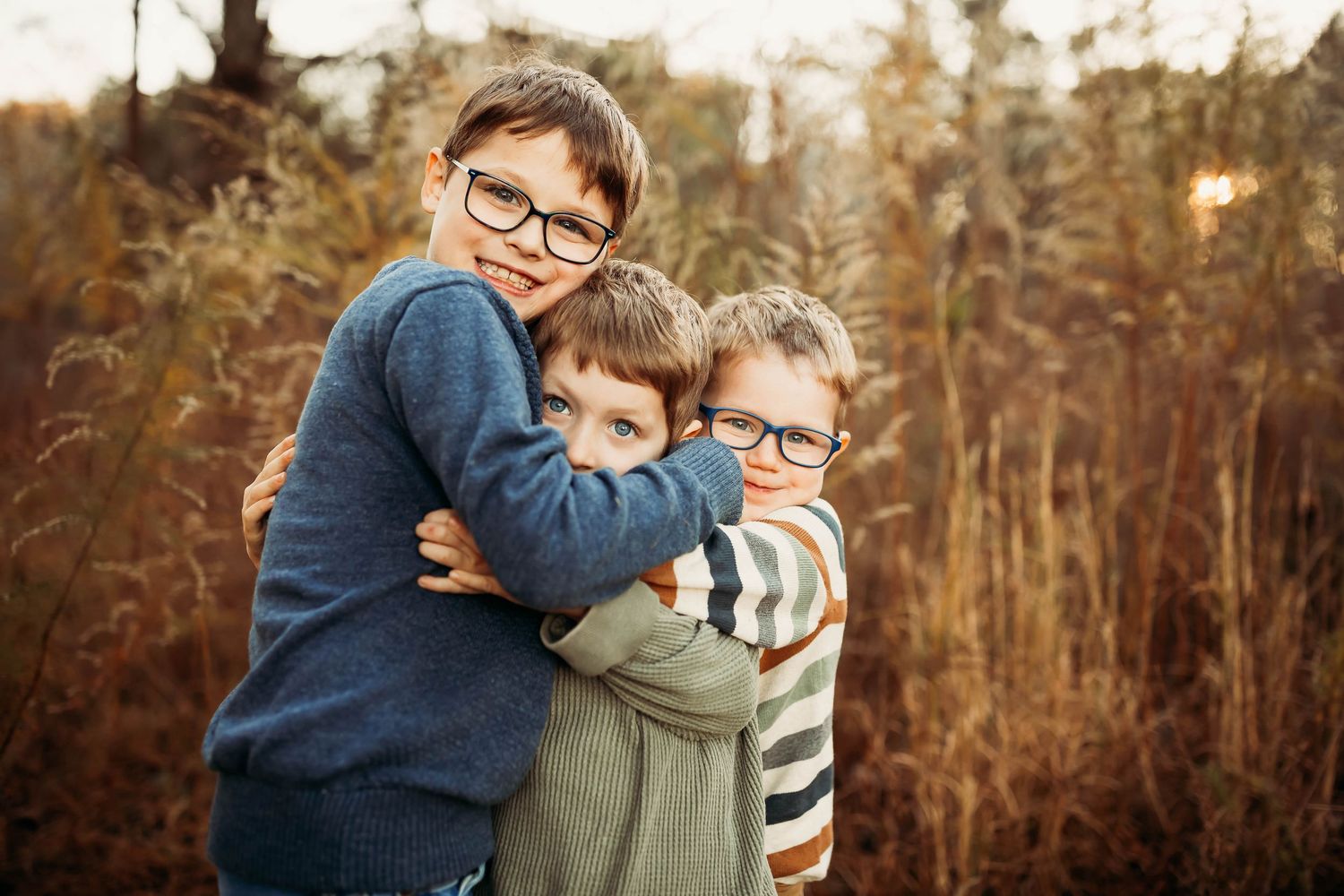 Three young boys wearing glasses share a warm group hug outdoors among tall autumn grasses.