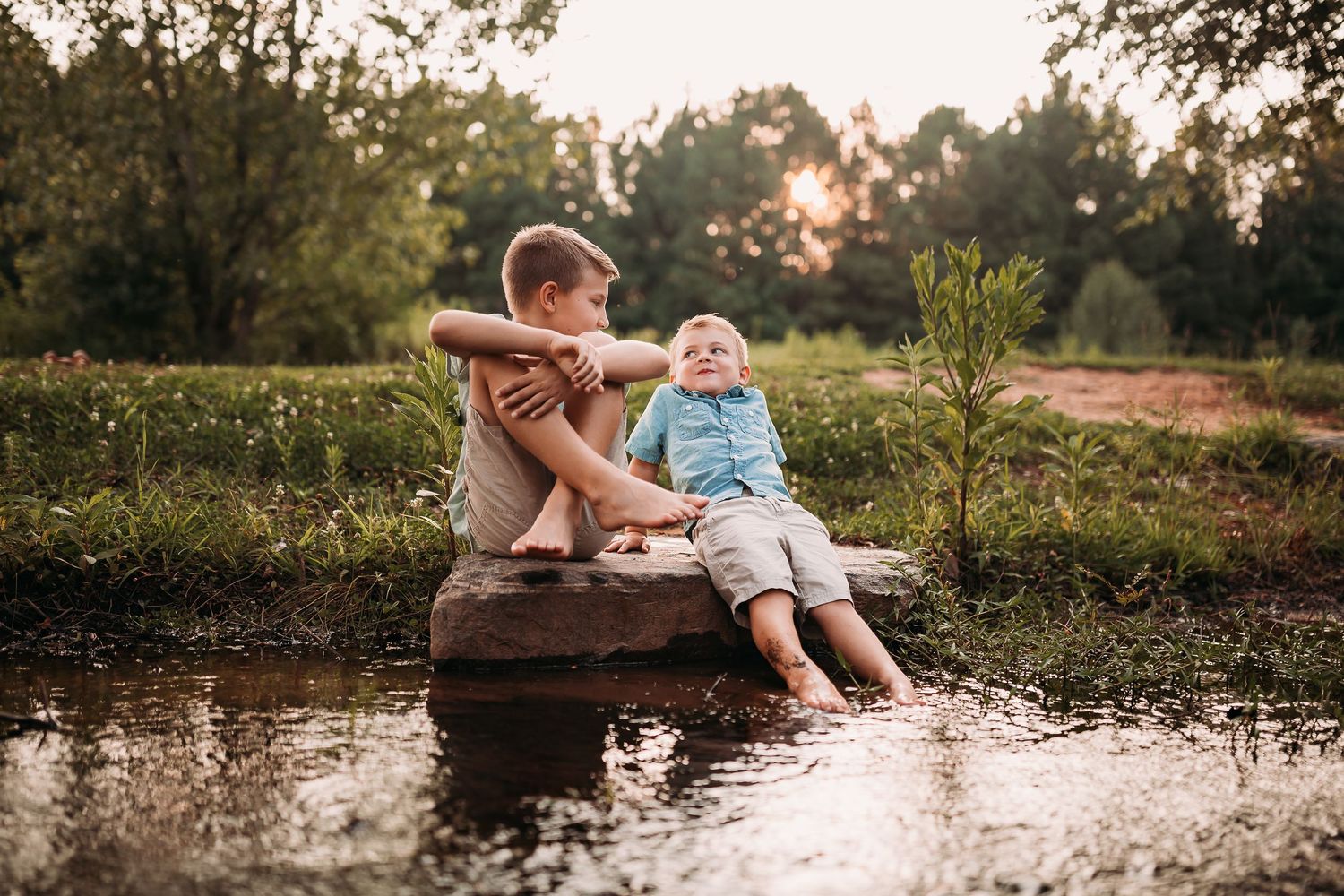 Two boys sitting on a rock by a peaceful pond, dangling feet in water, surrounded by lush greenery at sunset.