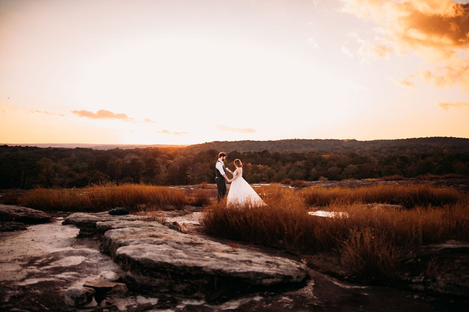 A bride and groom embrace on rocky terrain at sunset with a sweeping landscape and golden sky behind them.