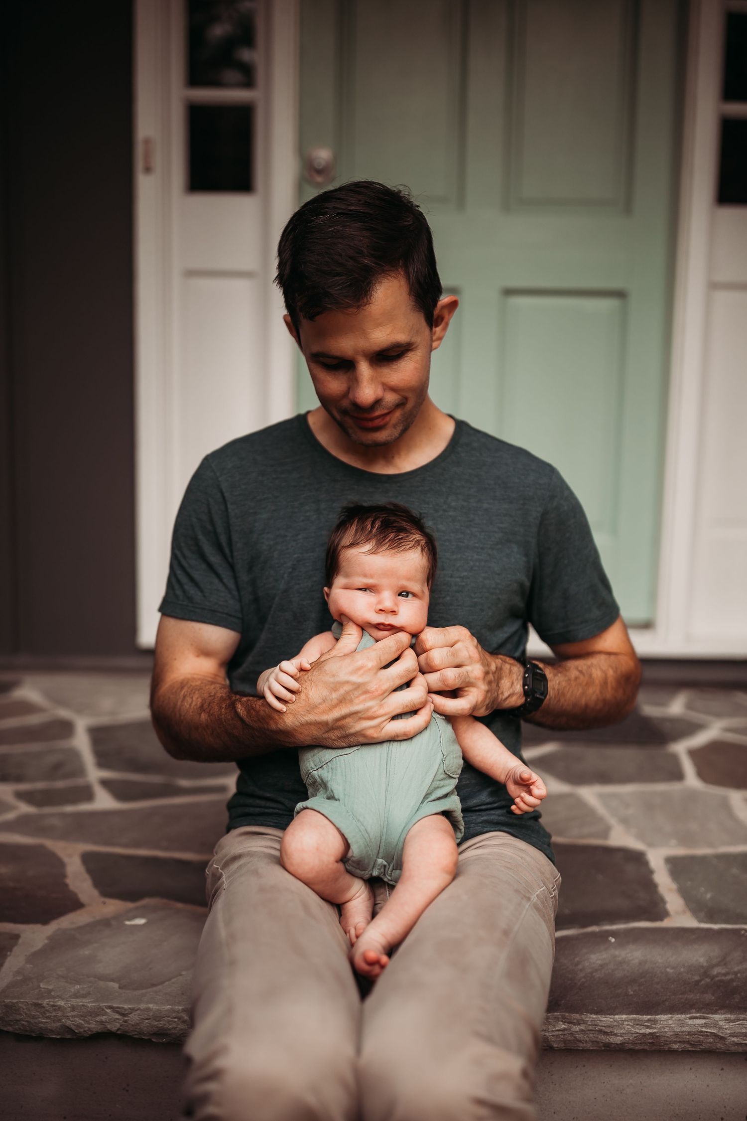 A father sits on stone steps holding his newborn baby in a light blue onesie outside a home.