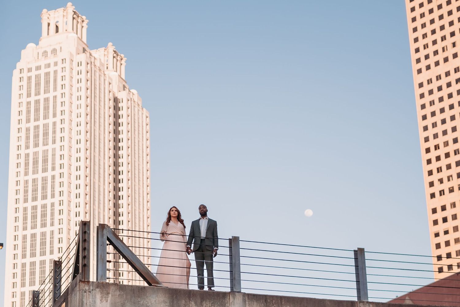 A couple stands on a rooftop with Atlanta skyscrapers and a full moon visible in the background at dusk.