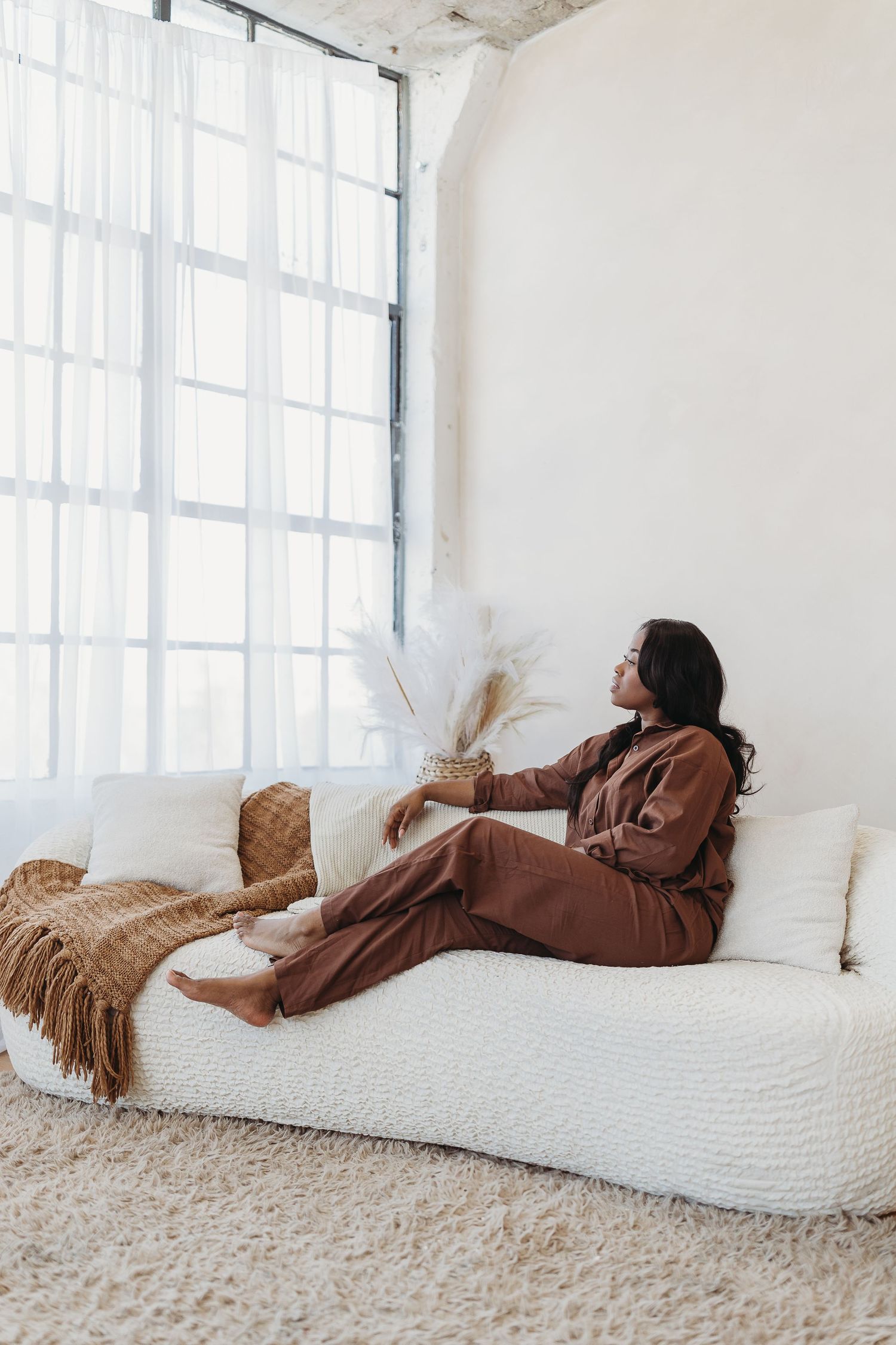 Woman in brown jumpsuit relaxing on white sofa near large windows in bright studio space.