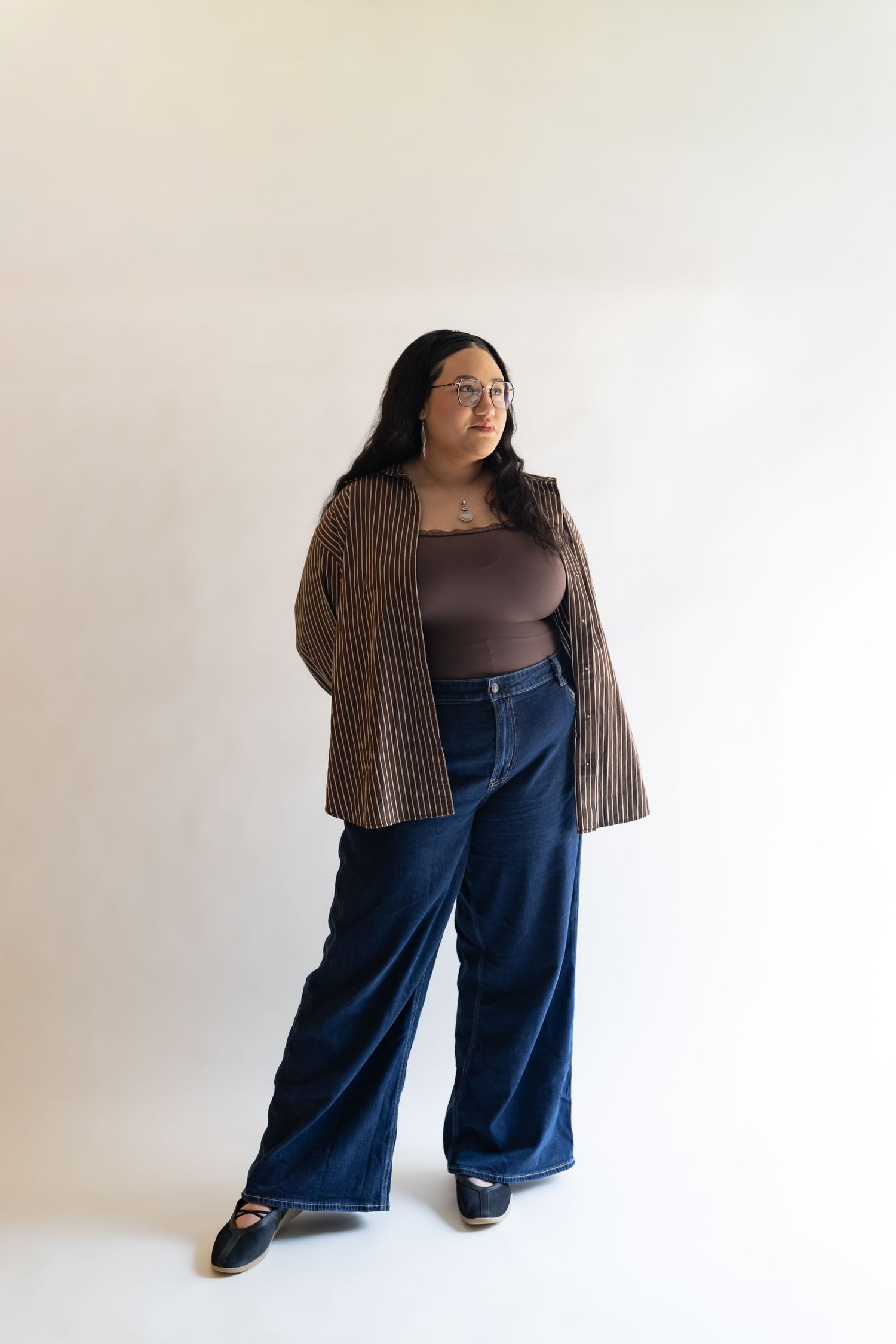 Woman in wide-leg jeans, brown bodysuit, striped cardigan, and black heels poses against white background.