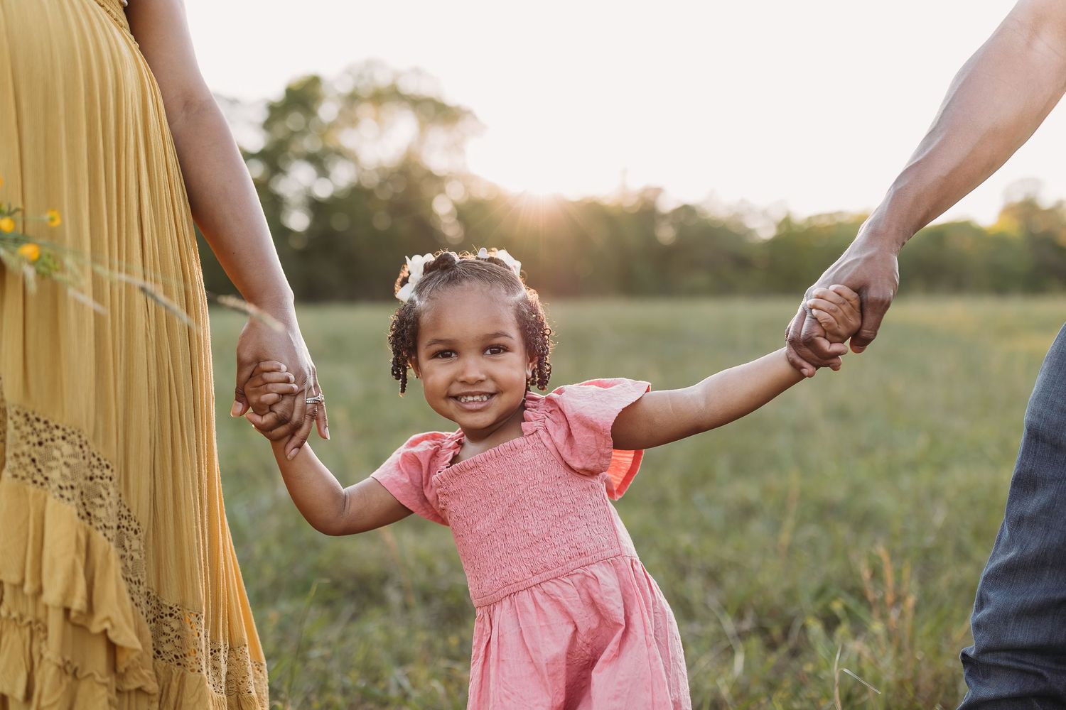 Young girl in pink dress smiles holding parents hands in sunny field at golden hour.