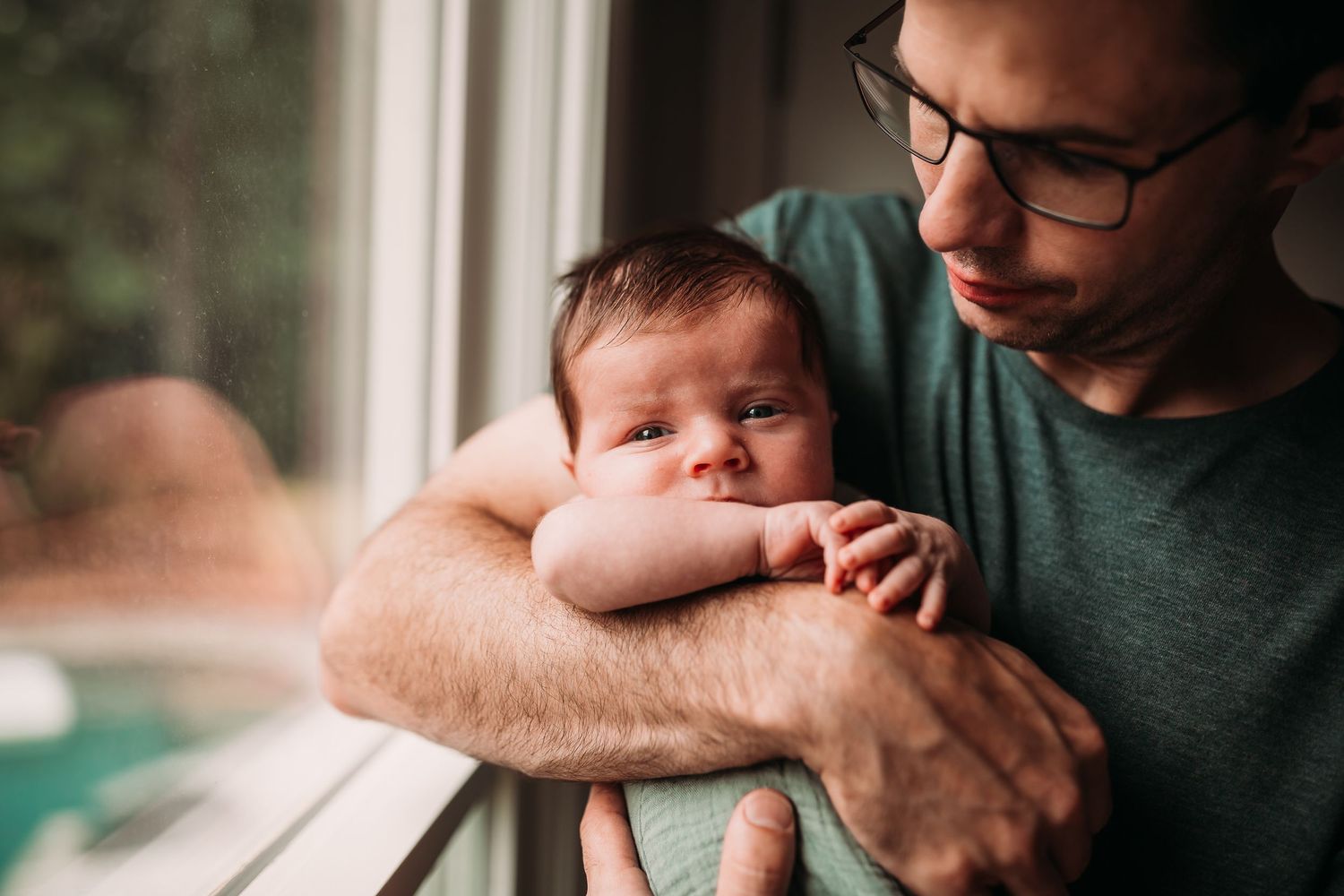 A father in glasses tenderly holds his newborn baby near a window in a heartwarming portrait.