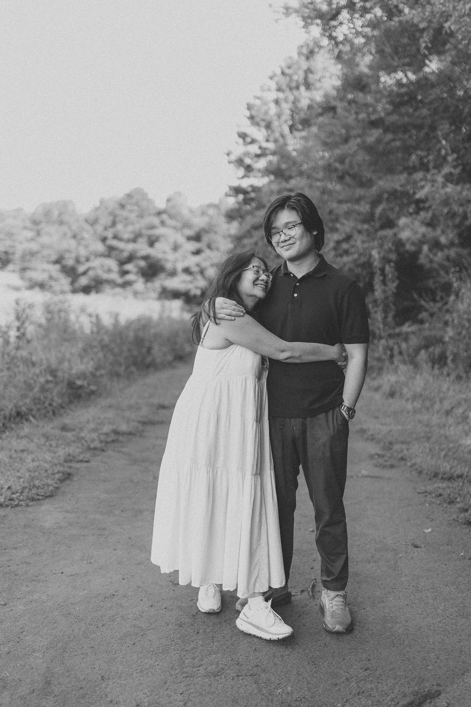 Black and white photo of a couple embracing on a scenic path surrounded by lush trees.