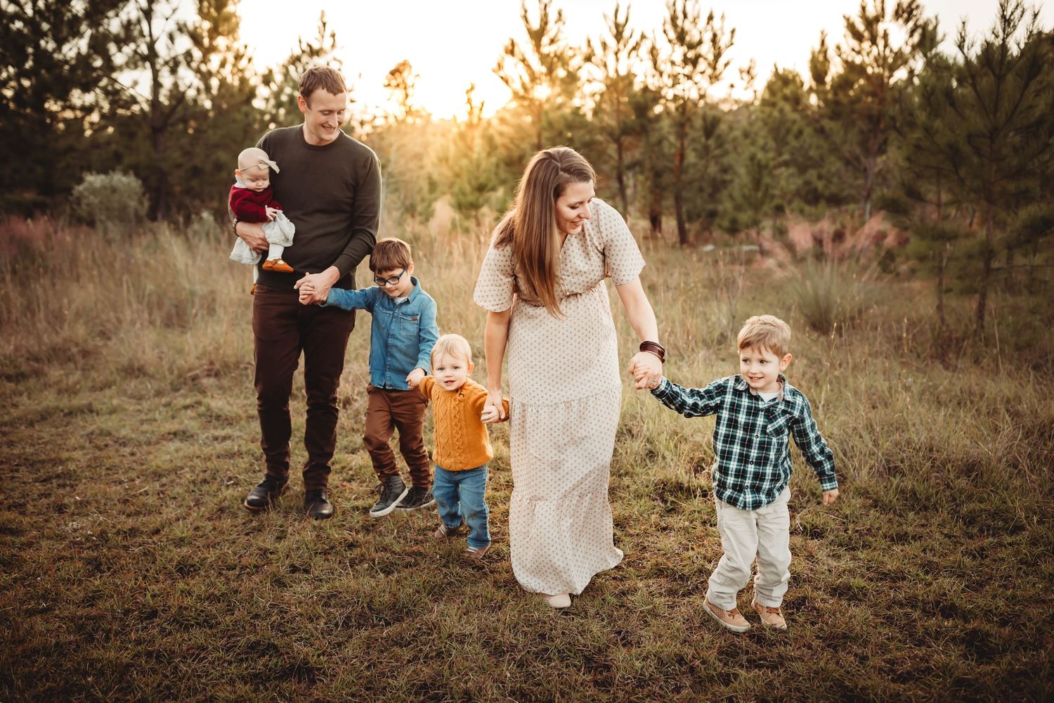 Family of five walking through a golden sunlit field at sunset, parents holding hands with young children in casual outdoor attire.