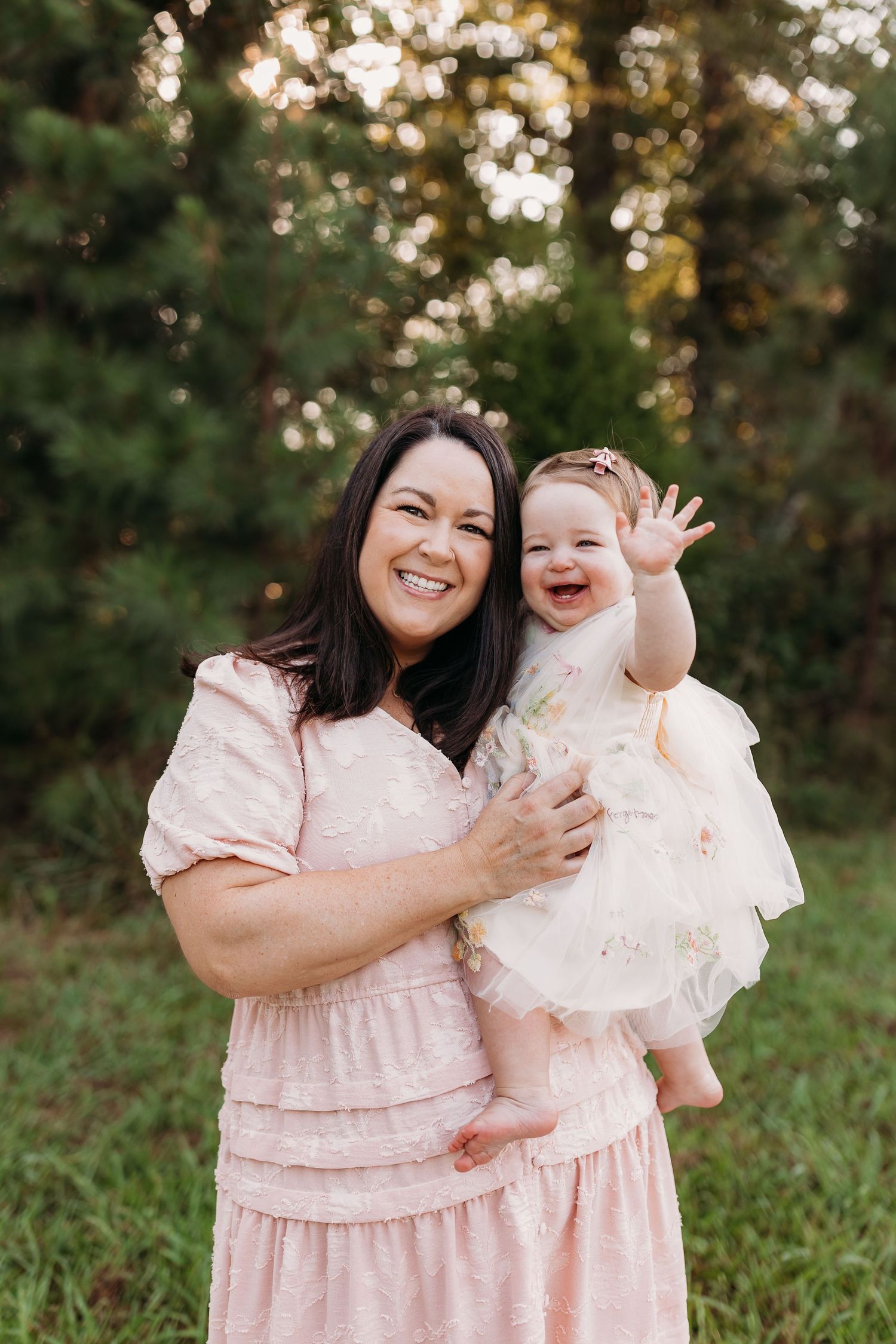 A smiling woman in a pink dress holds a laughing baby girl in a white dress outdoors near green trees.
