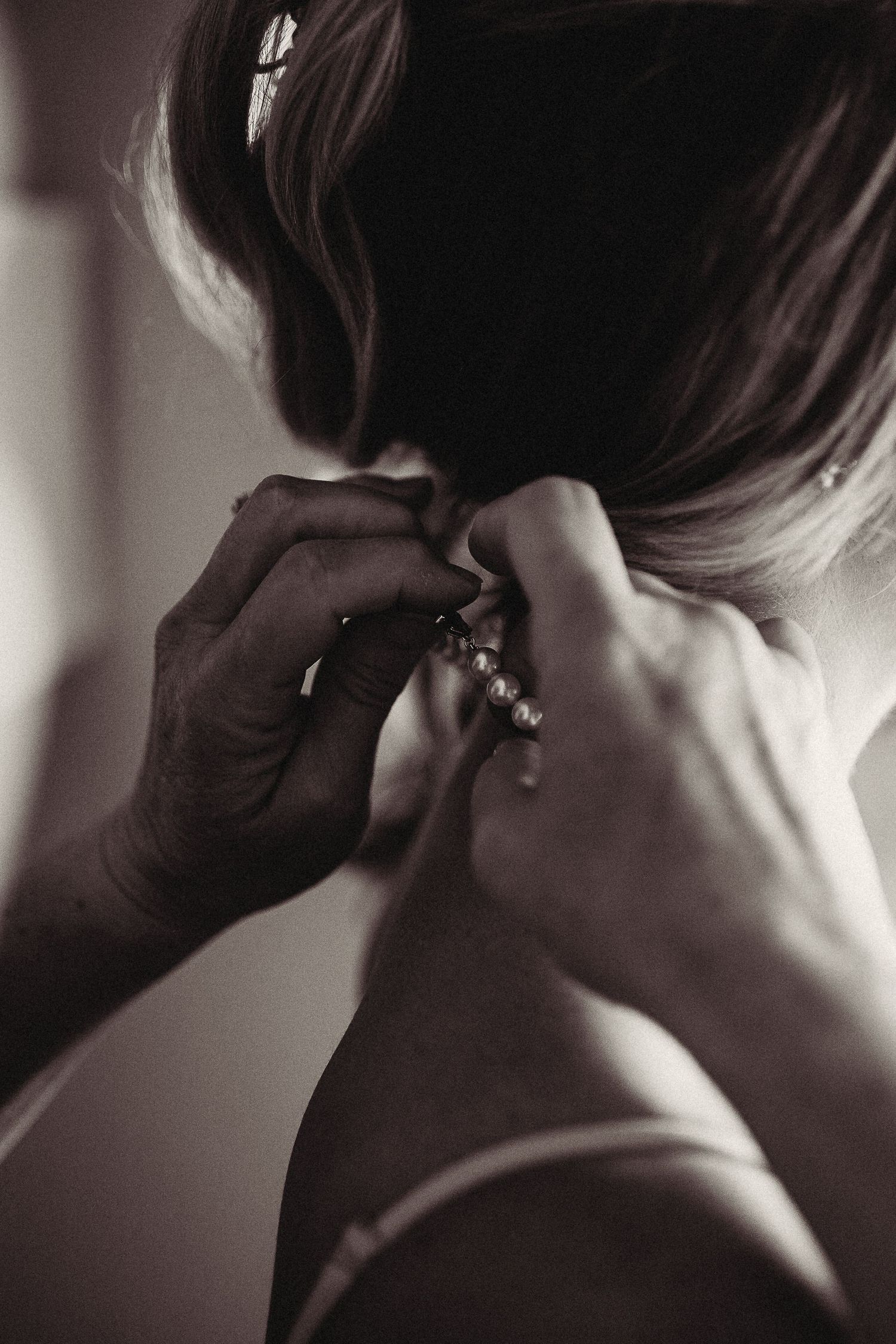 Black and white close-up of hands fastening a pearl necklace on a woman from behind.