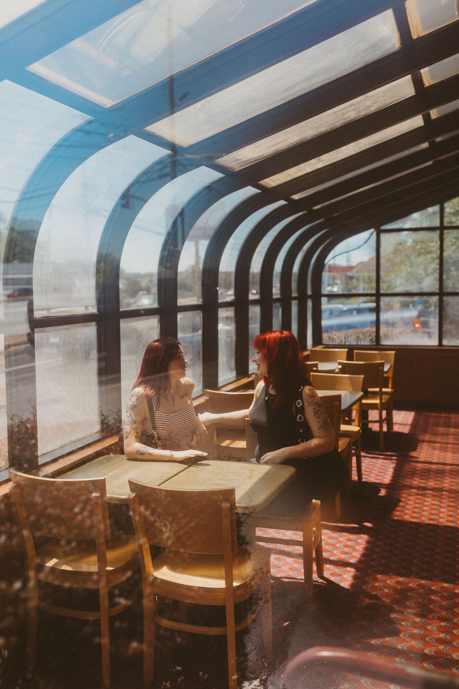 Two red-haired women conversing inside a sunlit glass-roofed diner booth, shot through the window.