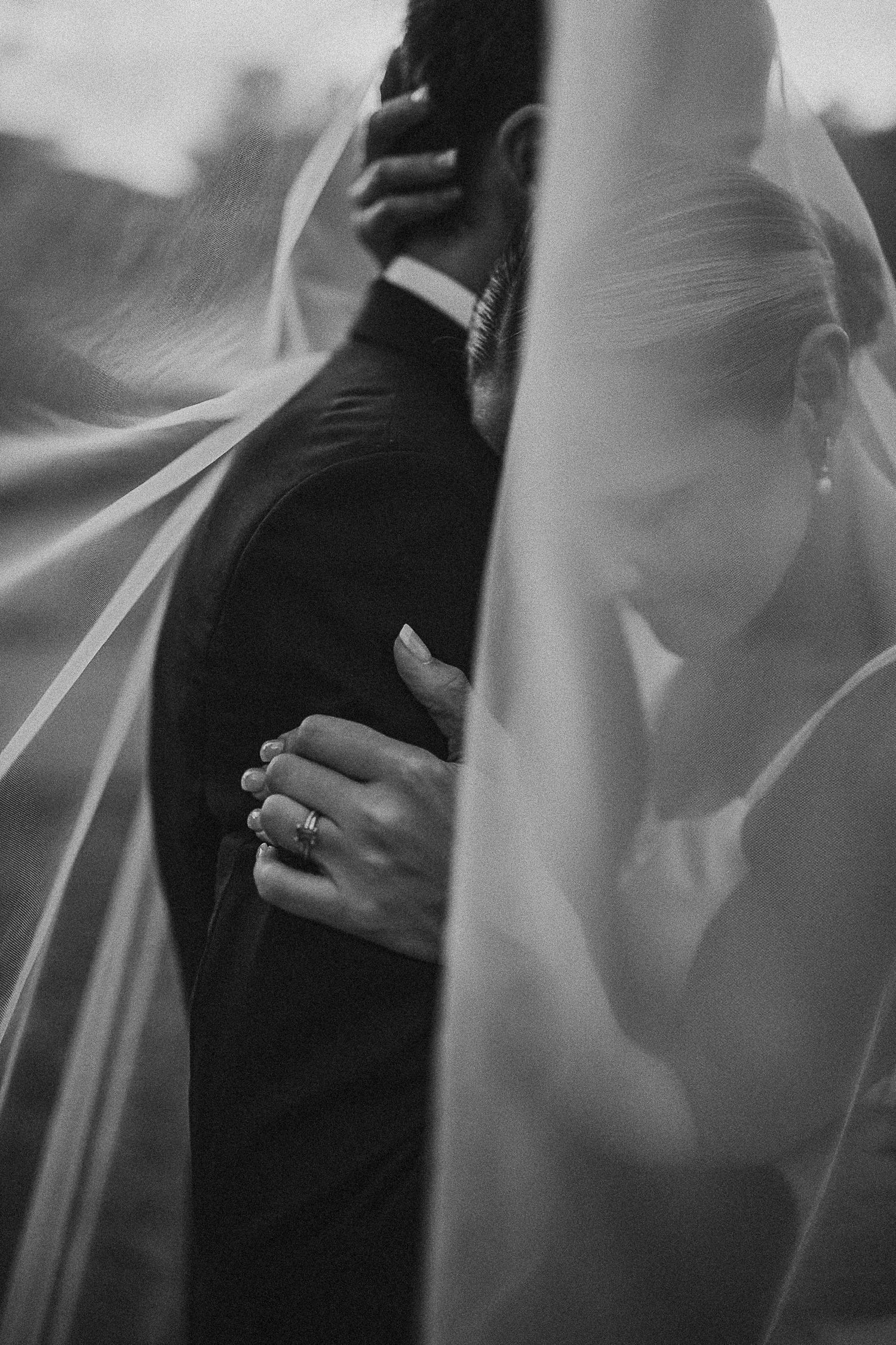 Intimate black and white wedding photo of bride and groom embracing beneath a flowing veil.