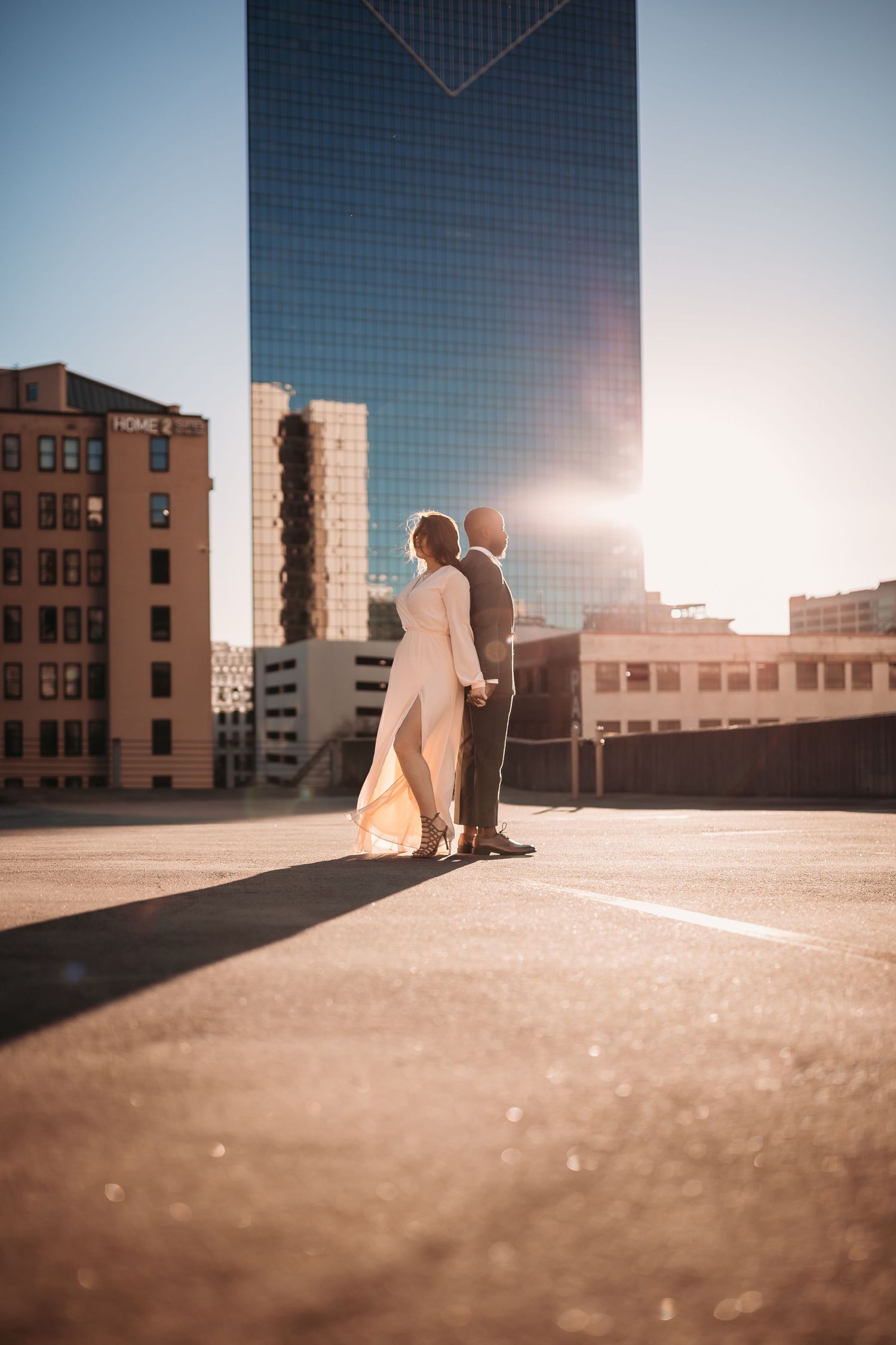 A couple in wedding attire stands on a rooftop parking lot with a glass skyscraper and sunburst behind them.
