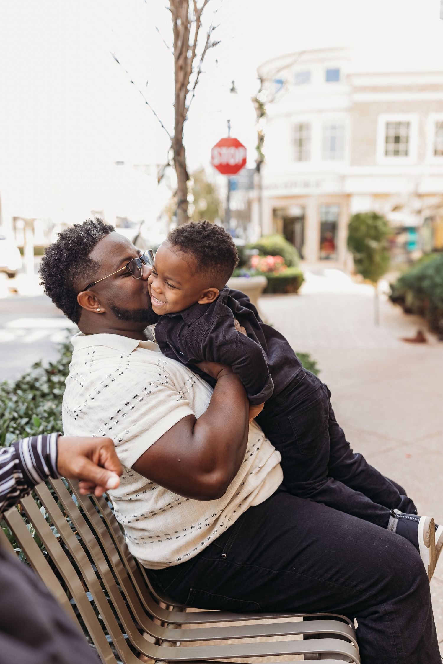 A father sits on a bench kissing his young son in an outdoor urban setting near a stop sign.
