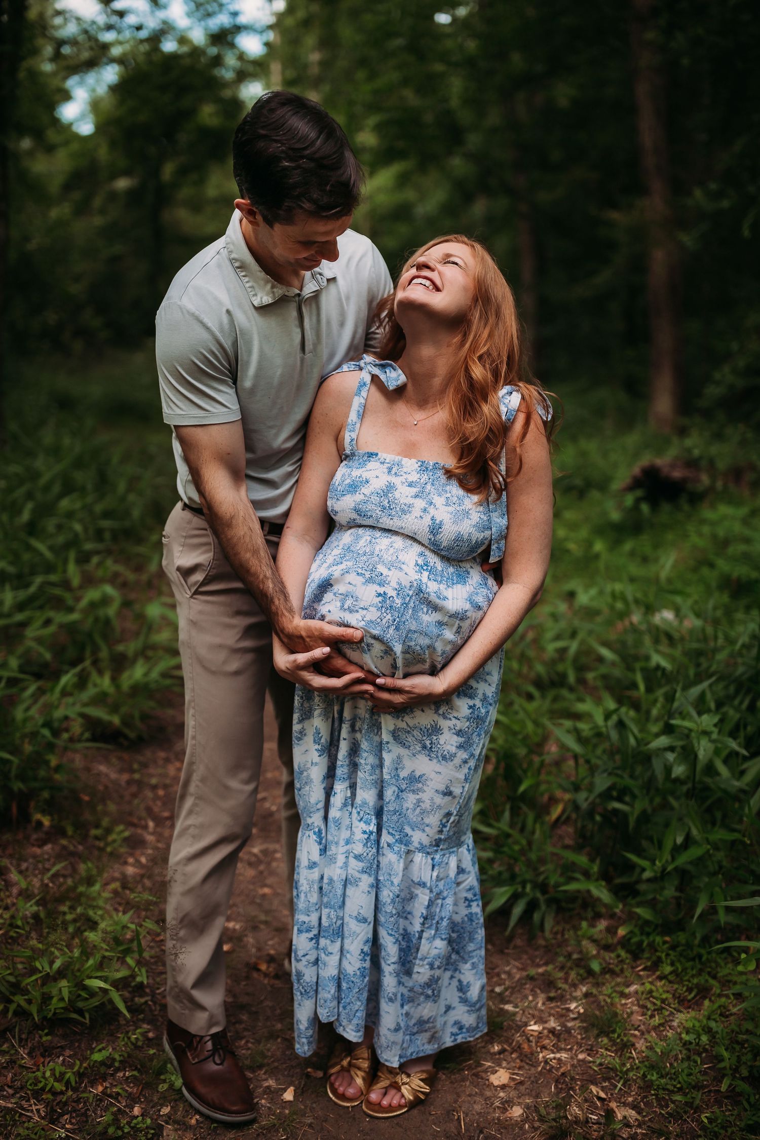 A joyful pregnant woman in a blue floral dress laughs with her partner outdoors in a lush green forest setting.
