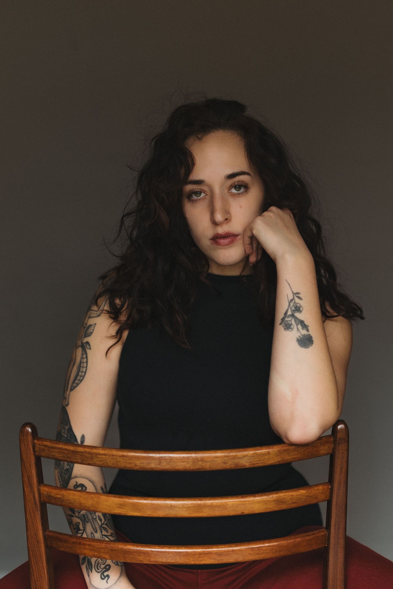 Tattooed woman with dark curly hair sitting on wooden chair, resting chin on hand.