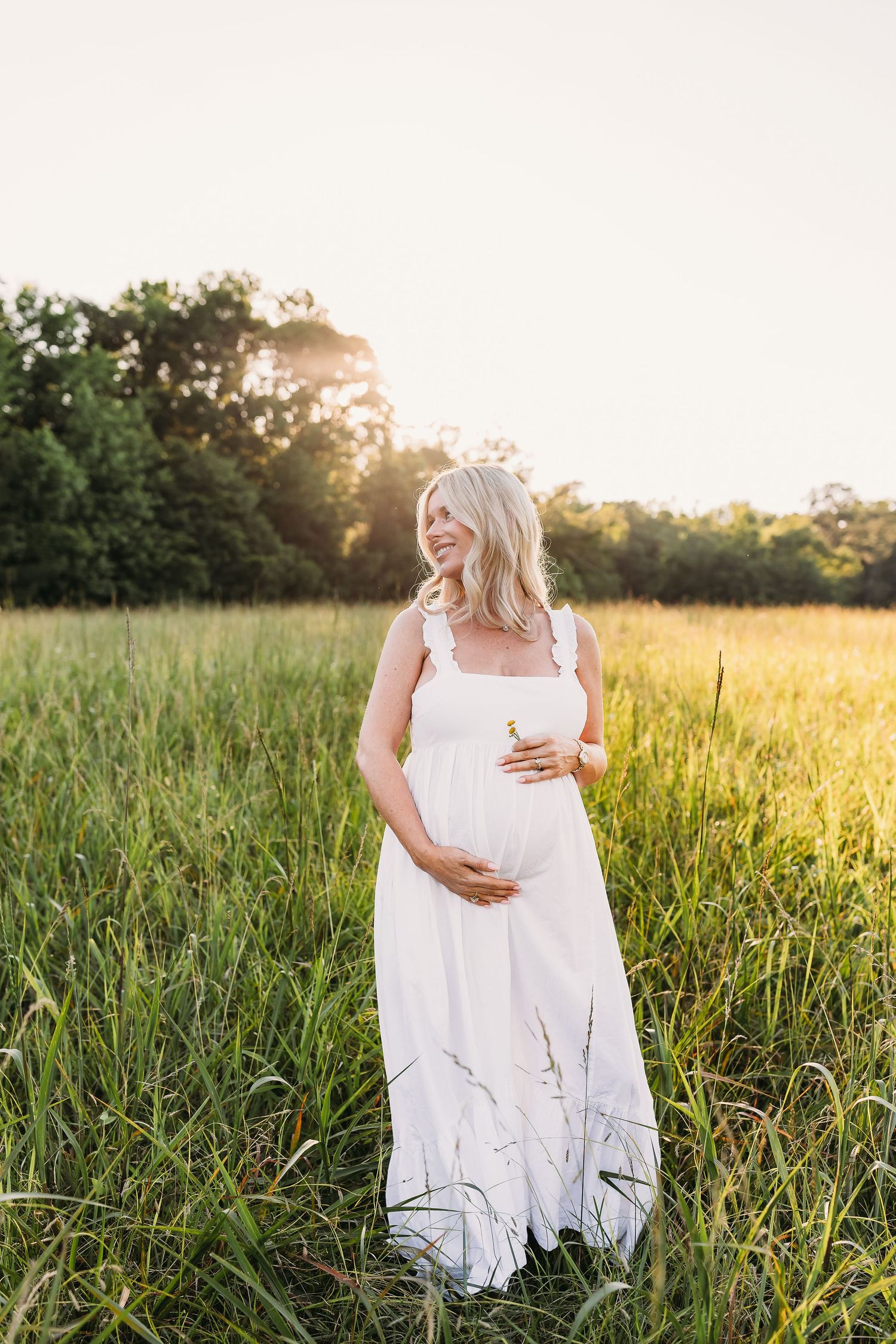 Smiling pregnant woman in white dress standing in sunny green field at golden hour, cradling baby bump.
