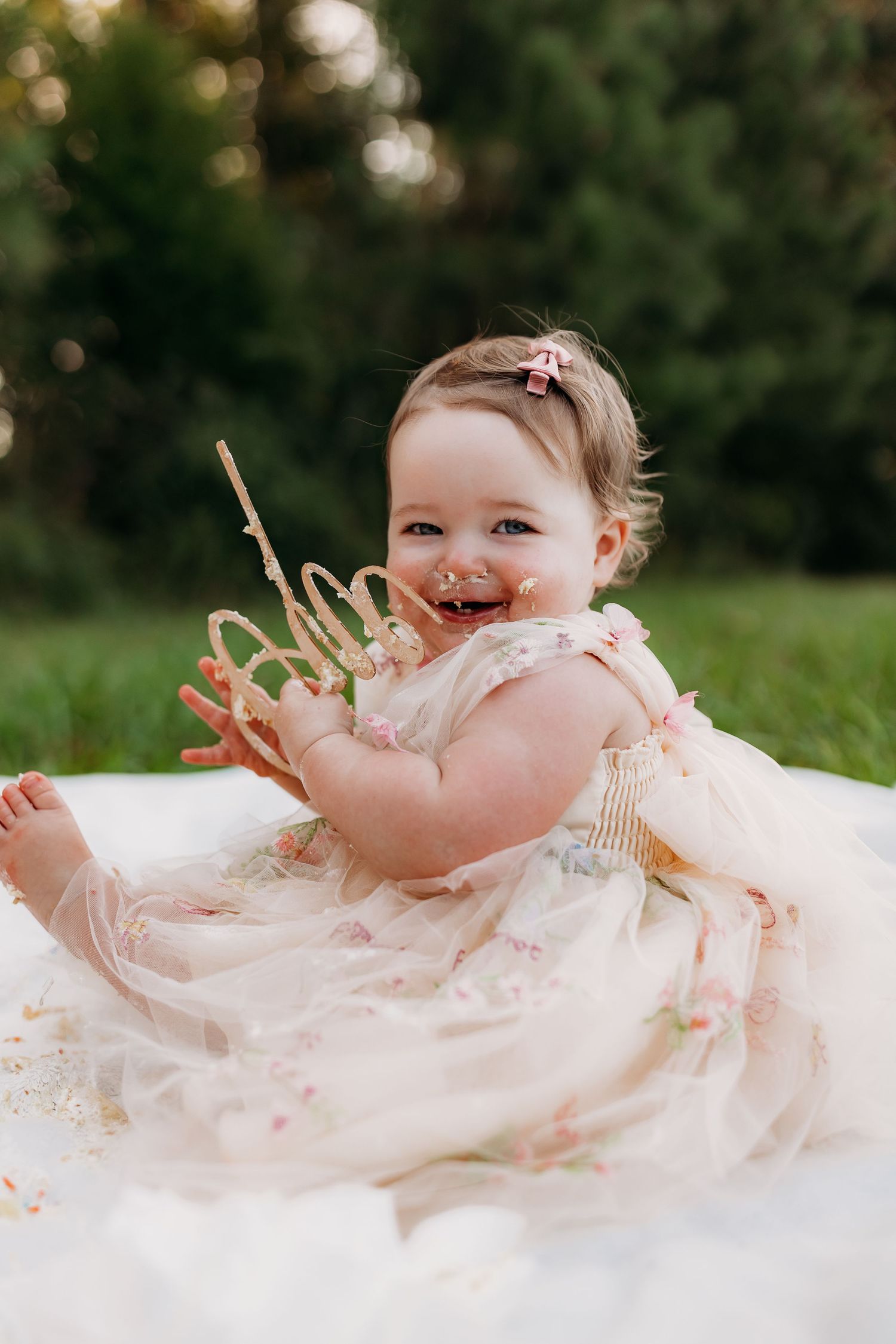 Smiling baby girl in floral dress holding birthday crown during outdoor cake smash photo session.