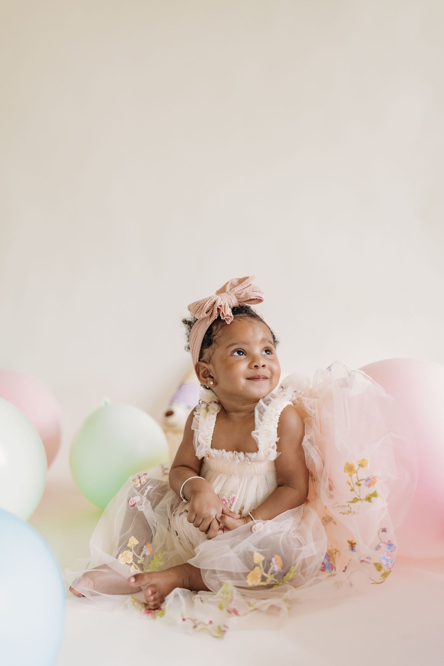 A smiling baby girl in a pink tulle dress and bow headband sits among pastel balloons during a birthday photo shoot.