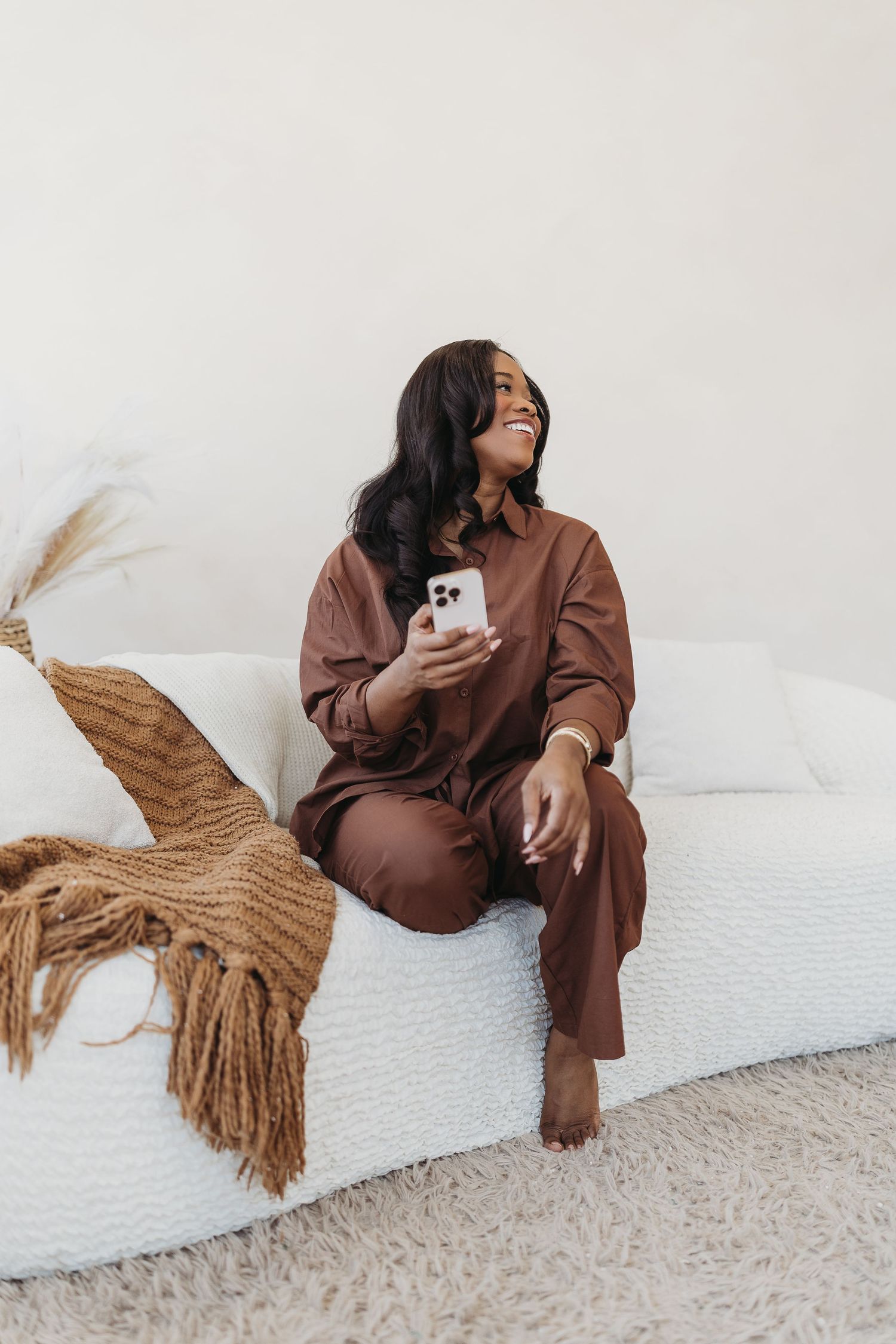 A happy woman in a brown outfit sits on a white sofa holding a smartphone in a bright, cozy room.