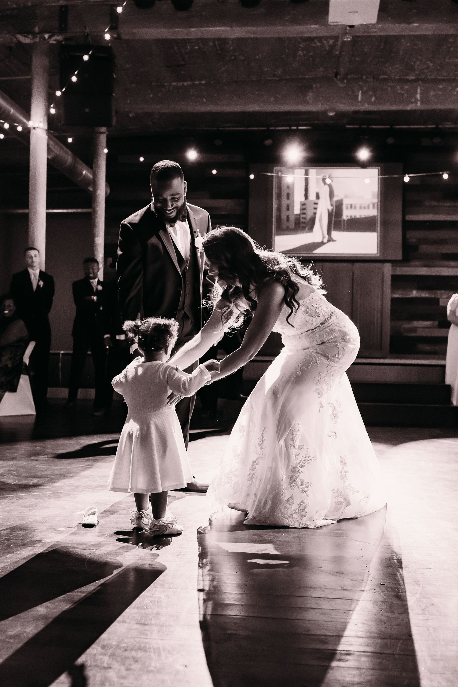 Bride and groom share a tender first dance moment with a flower girl on a black and white reception dance floor.