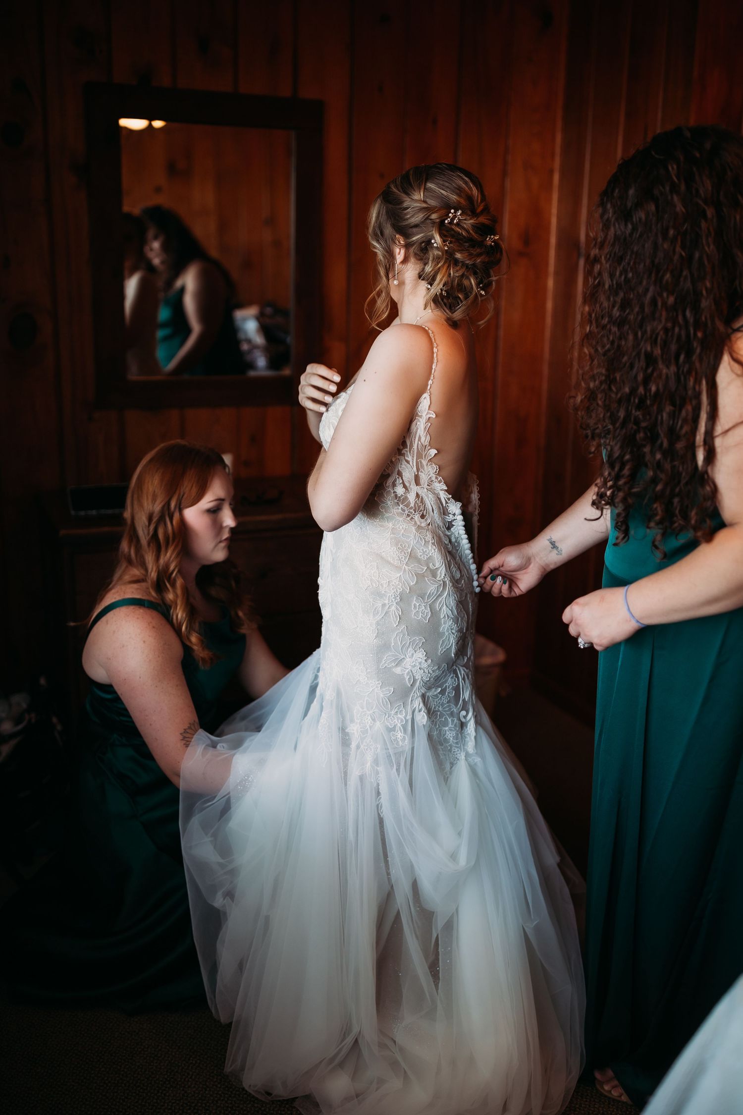 Bridesmaids in green dresses help bride into her lace wedding gown in a warmly lit wooden bridal suite.