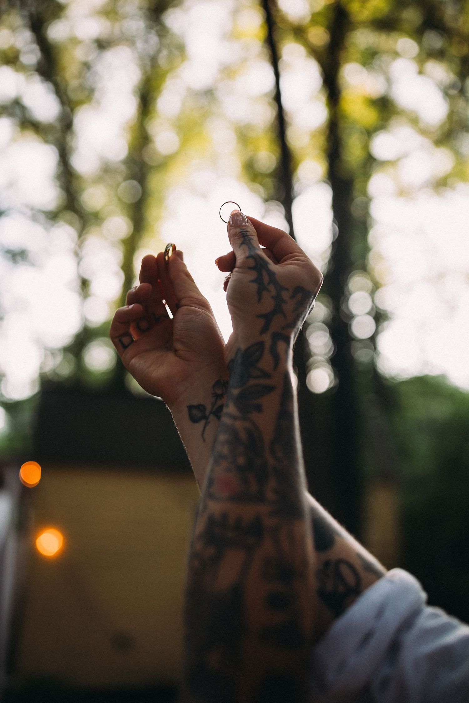 Tattooed hands raised holding wedding rings against a bokeh forest background at sunset.