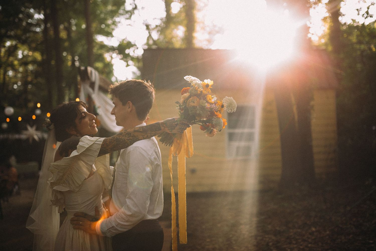 Couple embracing at golden hour in a forest, bride holding a floral bouquet with sunlight flare.