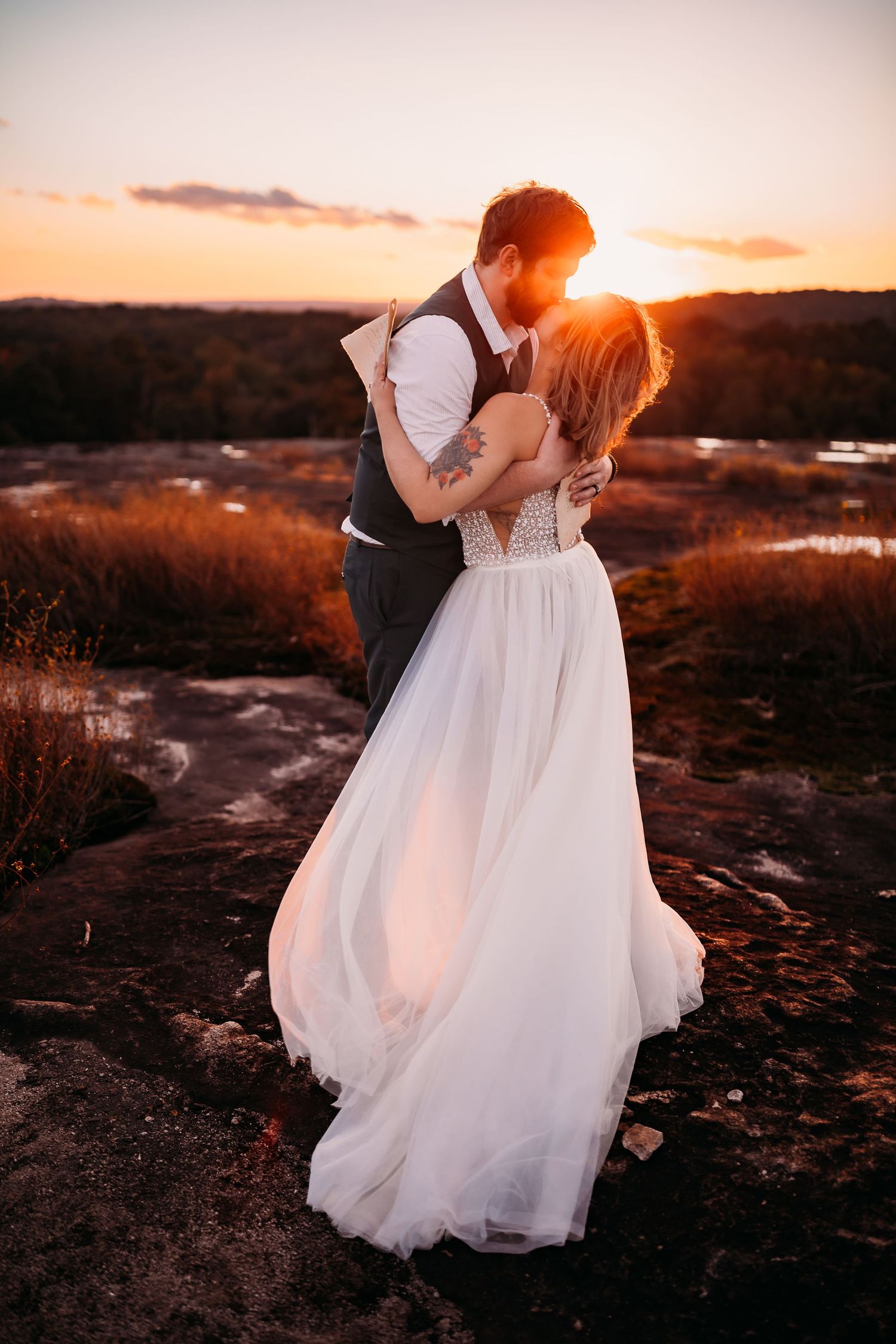 Newlyweds share a romantic kiss silhouetted against a golden sunset during their outdoor wedding portrait.