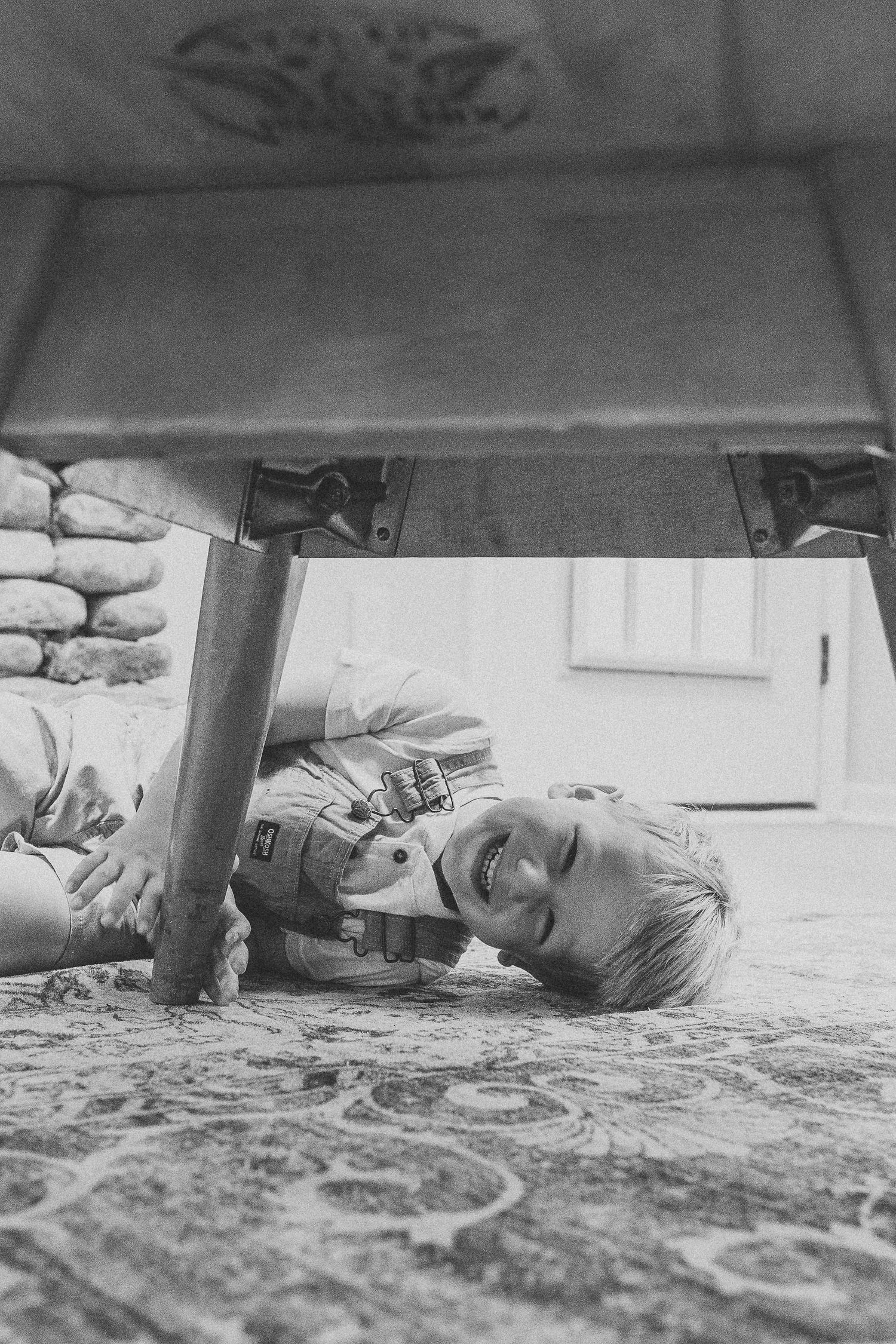 Black and white photo of a smiling child lying on a patterned carpet under a wooden table.