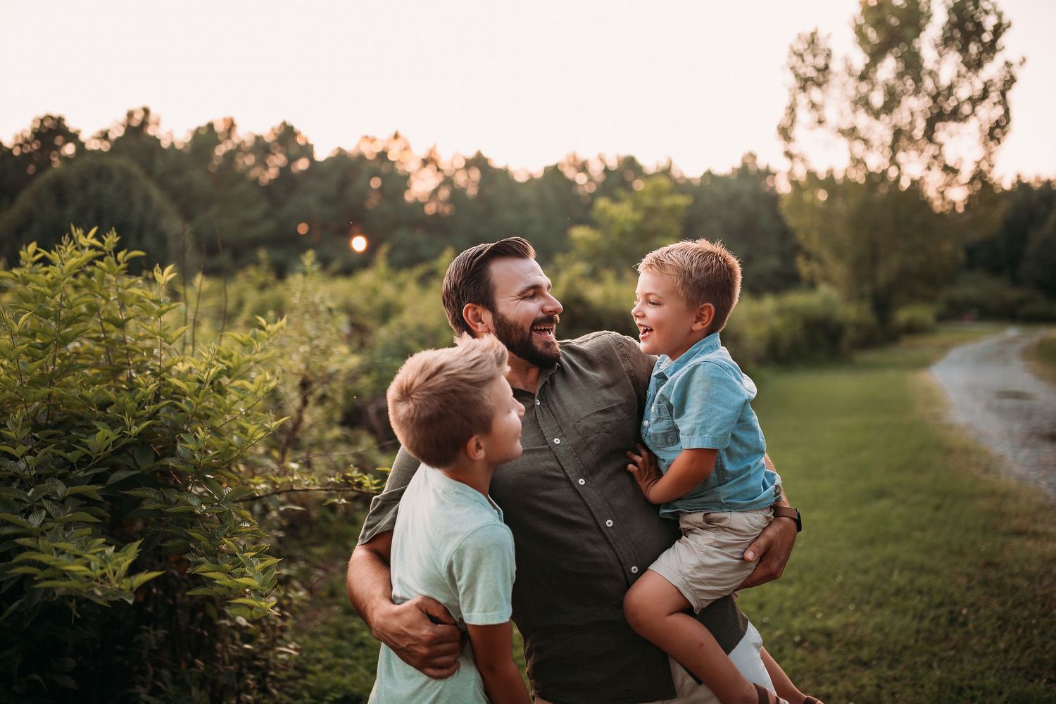 A father laughs joyfully with his two young sons outdoors at golden hour in a lush green field.