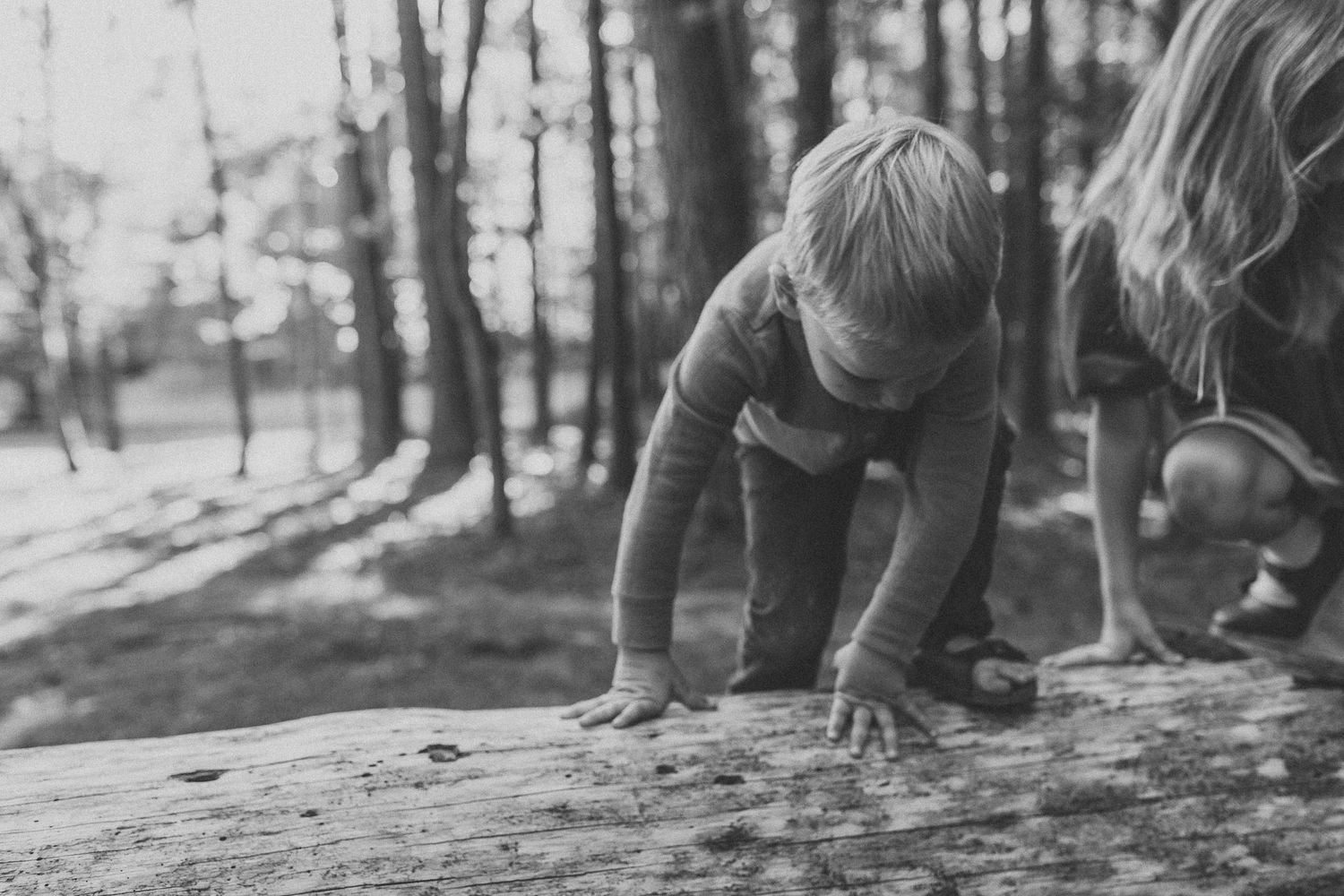 Two children climbing on a fallen log in a sunlit forest, captured in black and white photography.