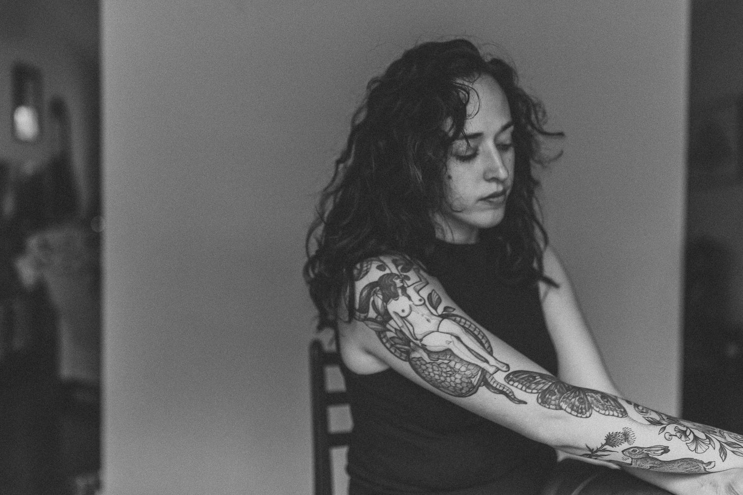 Black and white photo of a woman with curly hair and detailed sleeve tattoos looking downward pensively.
