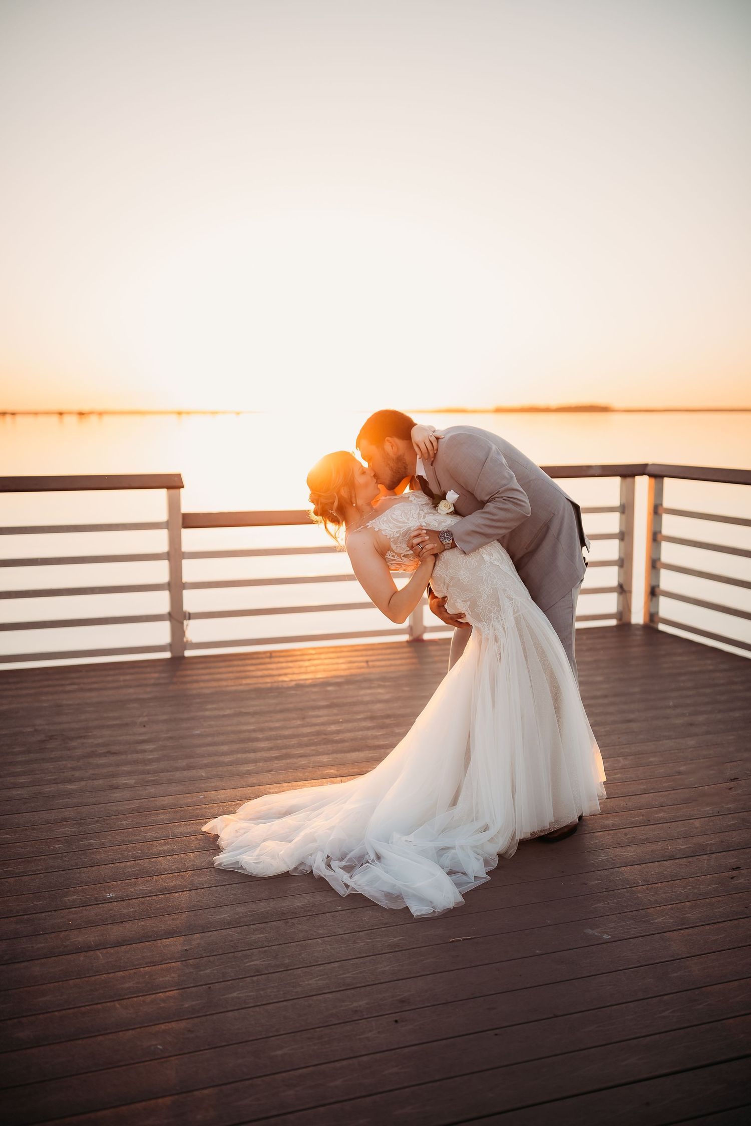 Groom dips bride in a romantic sunset kiss on a waterfront deck during their wedding portrait session.