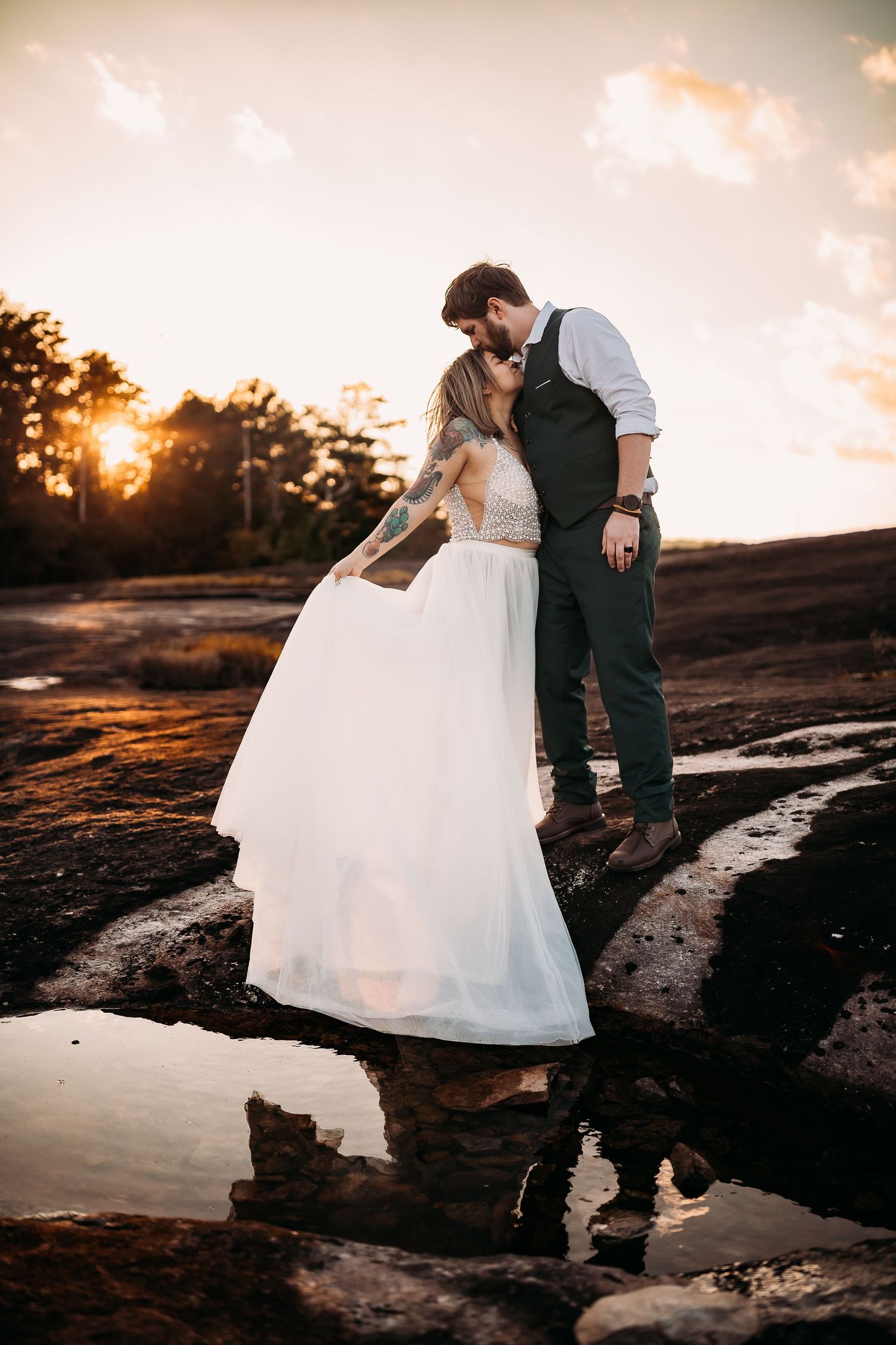 A bride and groom share a kiss on rocky terrain near a reflective puddle at golden hour sunset.