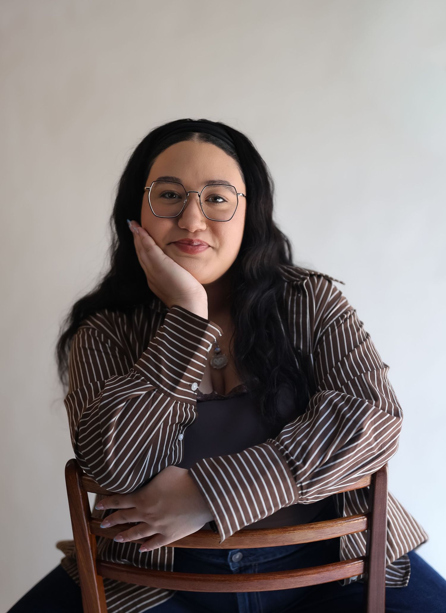 Young woman with long black hair and glasses sits relaxed on a wooden chair wearing a striped brown shirt.