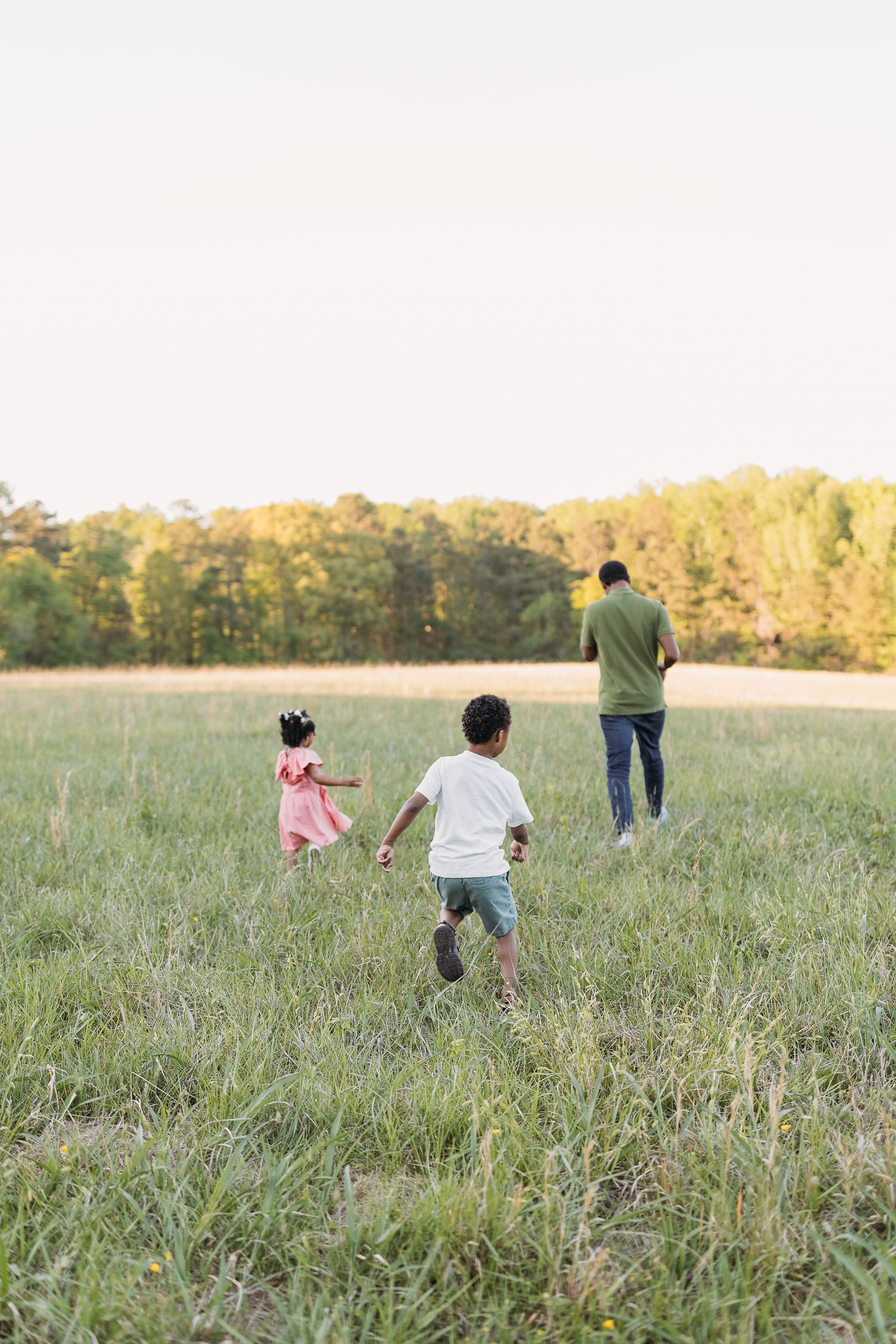 A family with two young children running through a green meadow toward a treeline at golden hour.