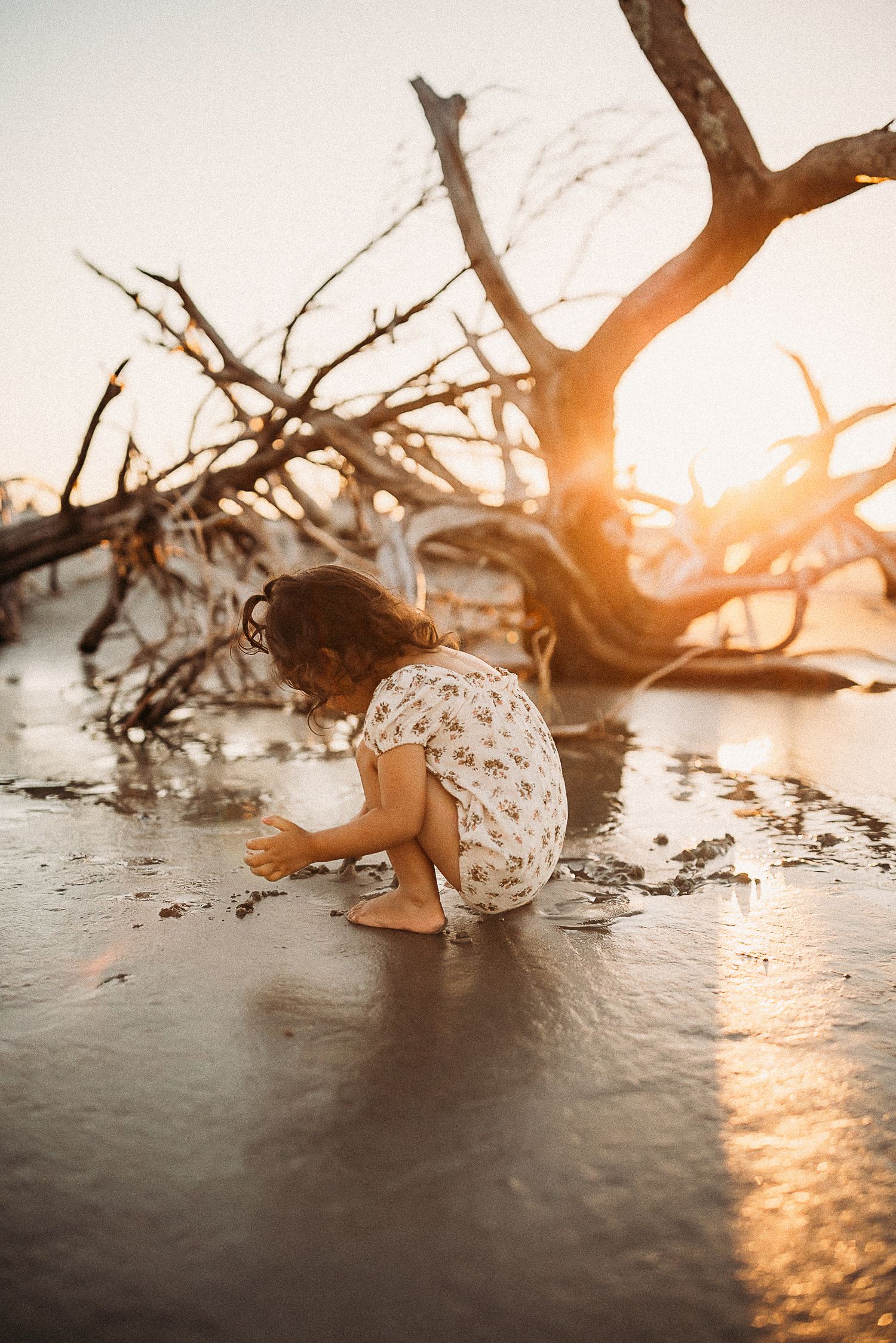 Young girl in floral dress crouching on wet beach sand at golden sunset near driftwood.