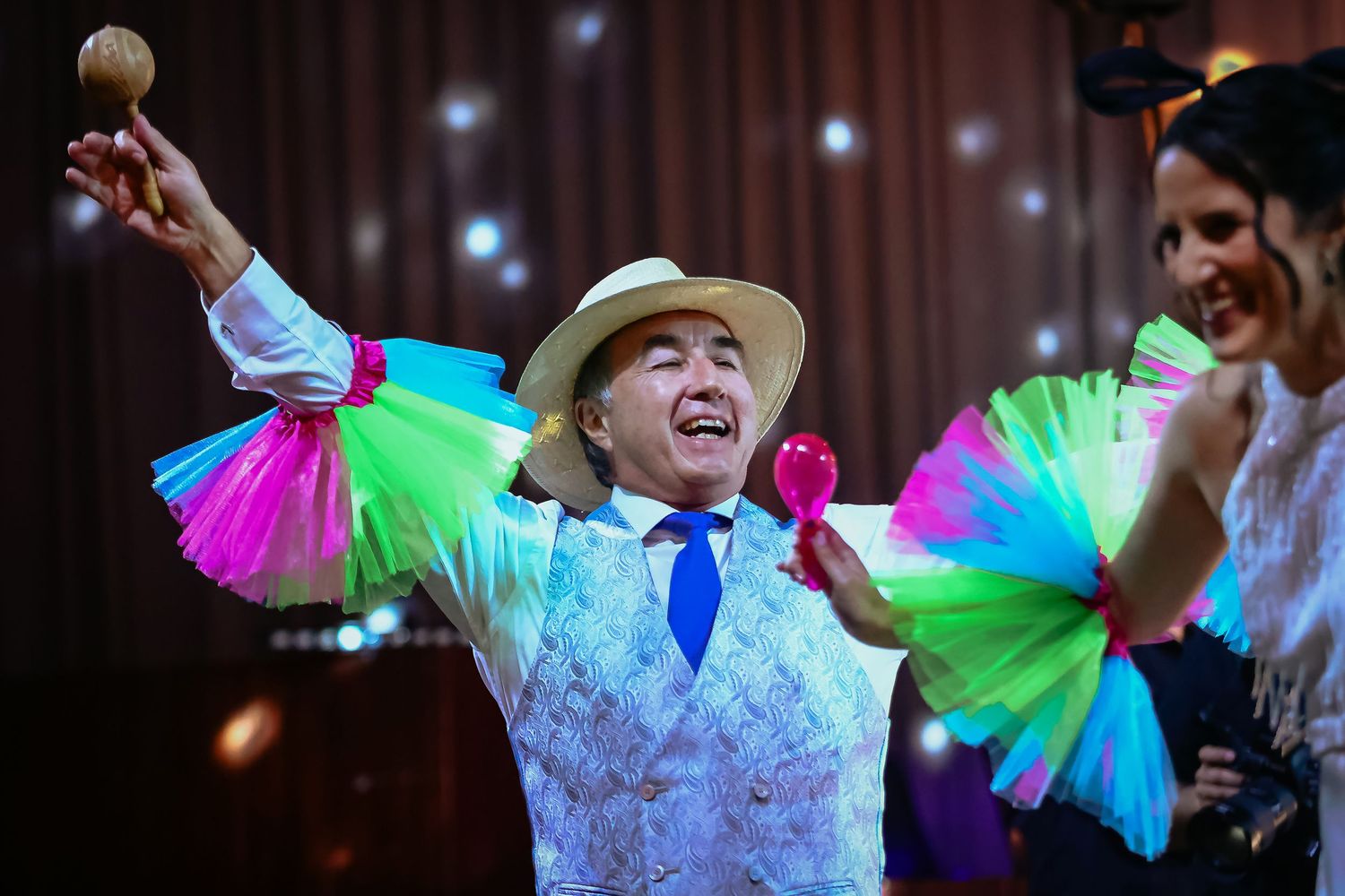 Festive performer in white hat dances with colorful neon fans and maracas at a lively celebration.