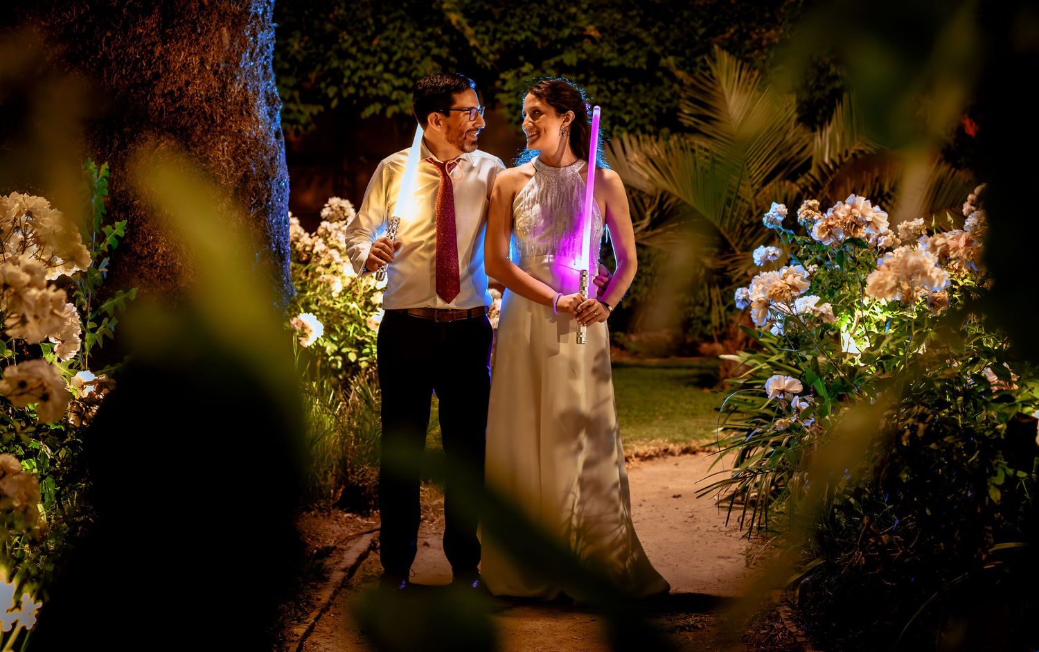 Couple shares a tender moment surrounded by flowers and glowing lightsabers at night.