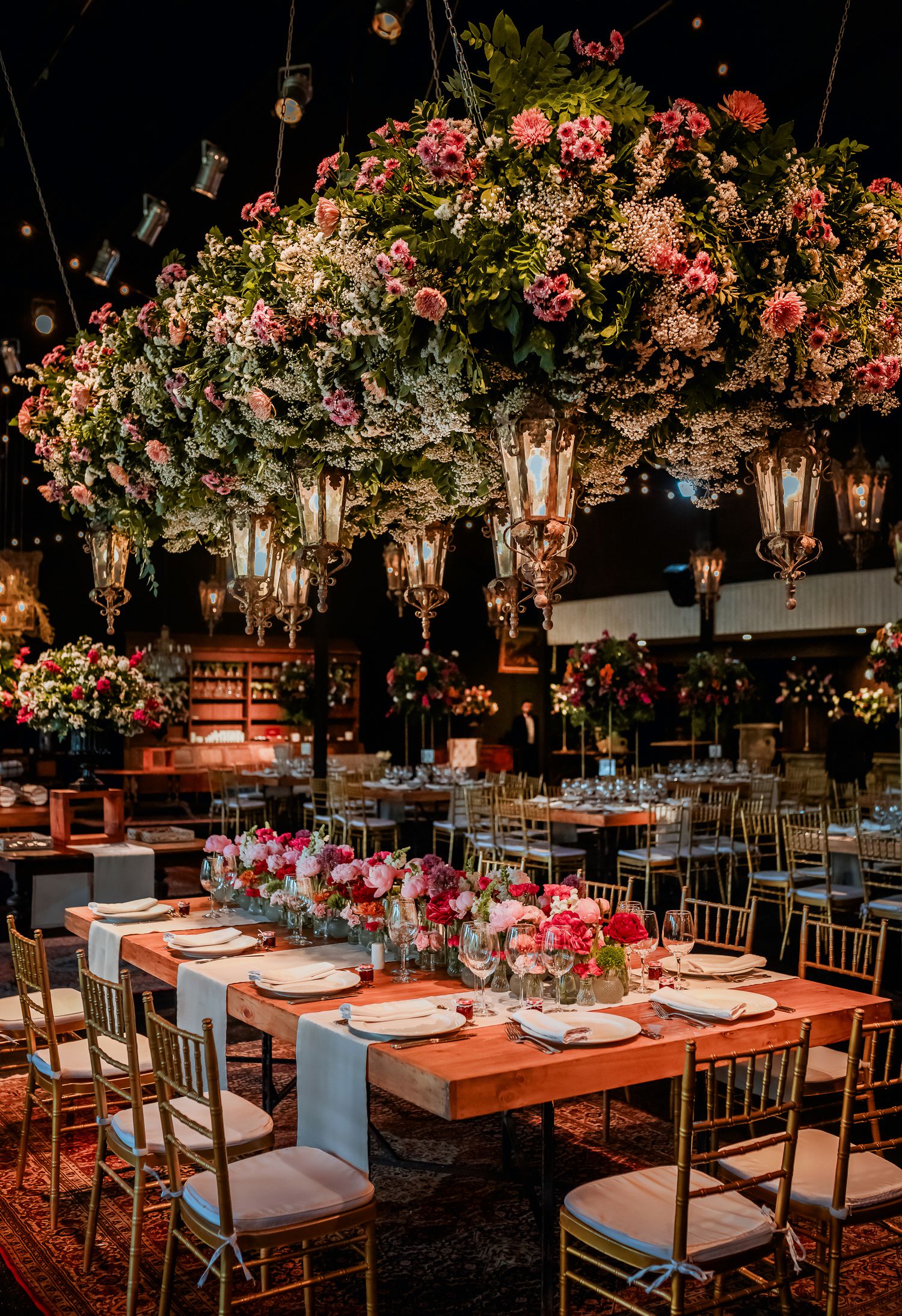 Elegant wedding reception with lush floral ceiling installation, lanterns, and wooden farm tables.