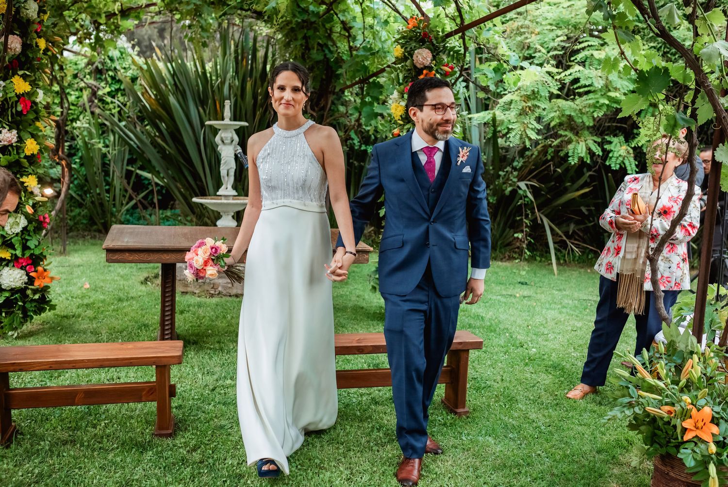 Bride and groom walk hand in hand down the aisle at an outdoor garden wedding ceremony.