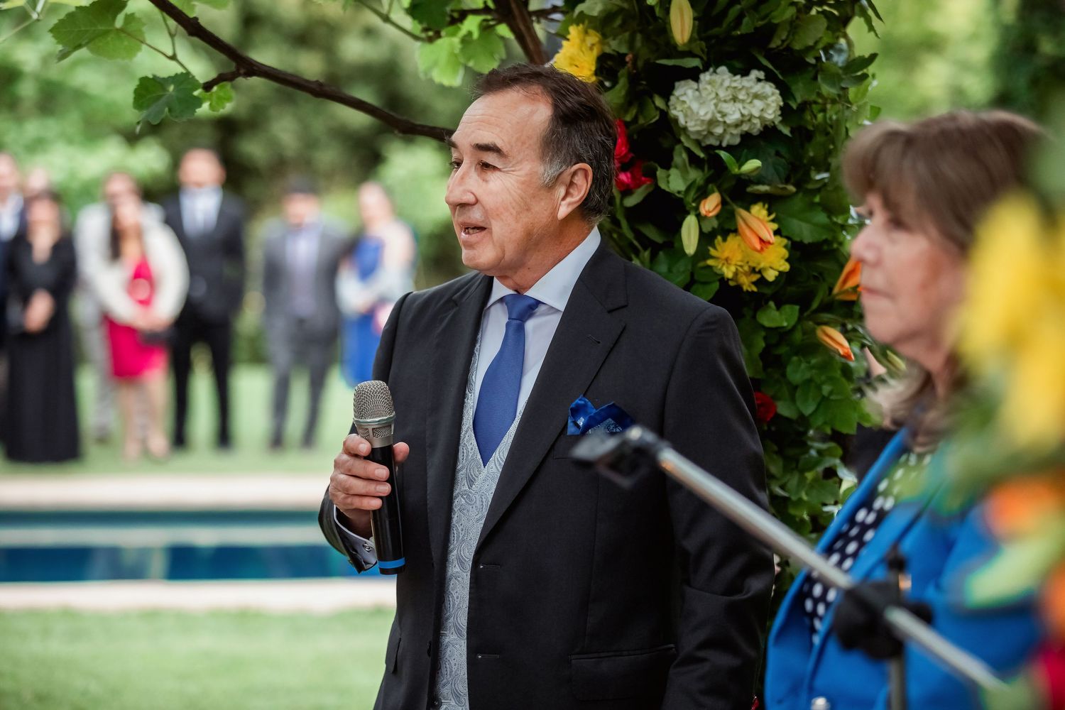 Older man in blue suit speaks at outdoor wedding ceremony with microphone.